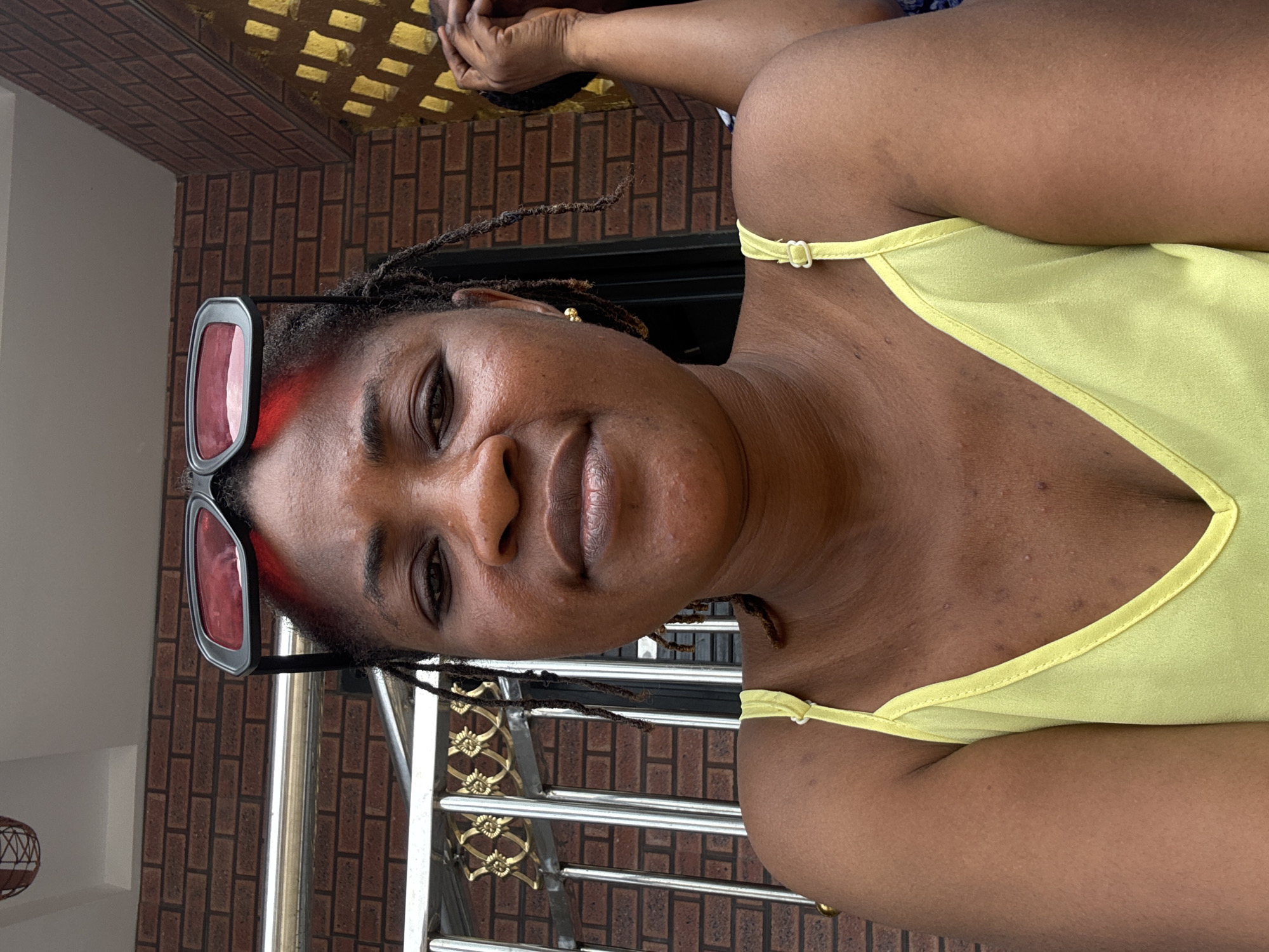 A medium shot captures a Black woman posing for a selfie outdoors in Ajara, Nigeria. She is positioned centrally in the frame, facing the camera directly. The woman has dark skin, dark hair styled into locs, and is wearing red-tinted sunglasses perched on top of her head. Her eyes are open and looking forward. She is dressed in a light yellow tank top with thin straps. Her left shoulder and upper chest are visible, revealing some skin texture. In the background, a brick wall is visible, along with a metal railing featuring decorative gold elements. To the right of the woman, another person's arm and hand are partially visible, with their hand touching their face. The overall environment appears to be outdoors, possibly a porch or entryway, with a hint of daylight.