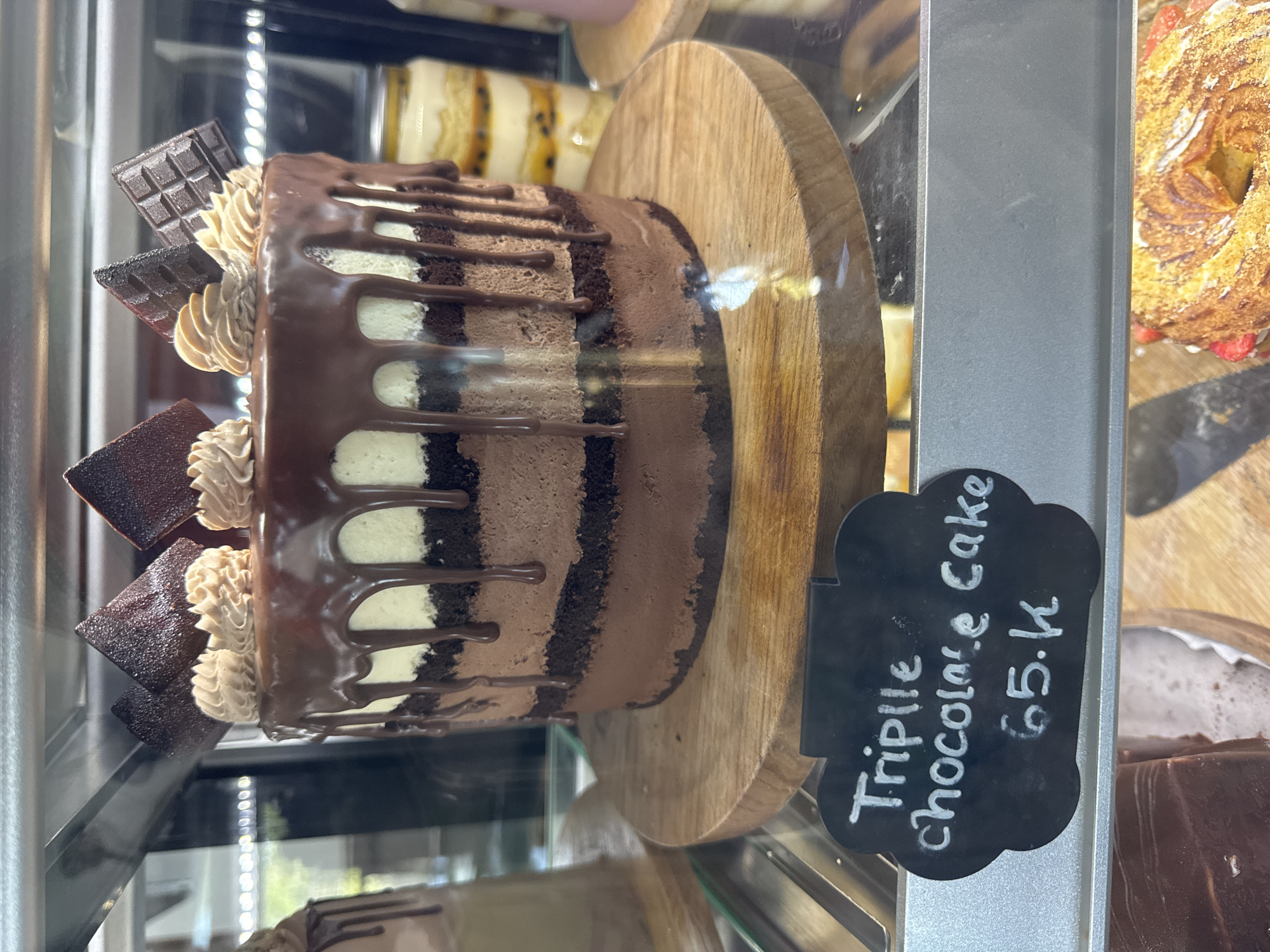 The image captures a slice of triple chocolate cake displayed in a glass case, likely in a bakery or cafe. The cake sits on a wooden serving board, accompanied by a small black signboard stating "Triple Chocolate Cake 65.k". The cake has three distinct chocolate layers with white cream filling. The top layer is coated with dark chocolate and decorated with chocolate pieces and swirls of beige icing. Drizzled chocolate accents adorn the cake. Other pastries and baked goods are visible in the background. It can be inferred from the price that the image was taken in Kuta Selatan, Indonesia where it is common to denominate prices by 1000's or "k".
