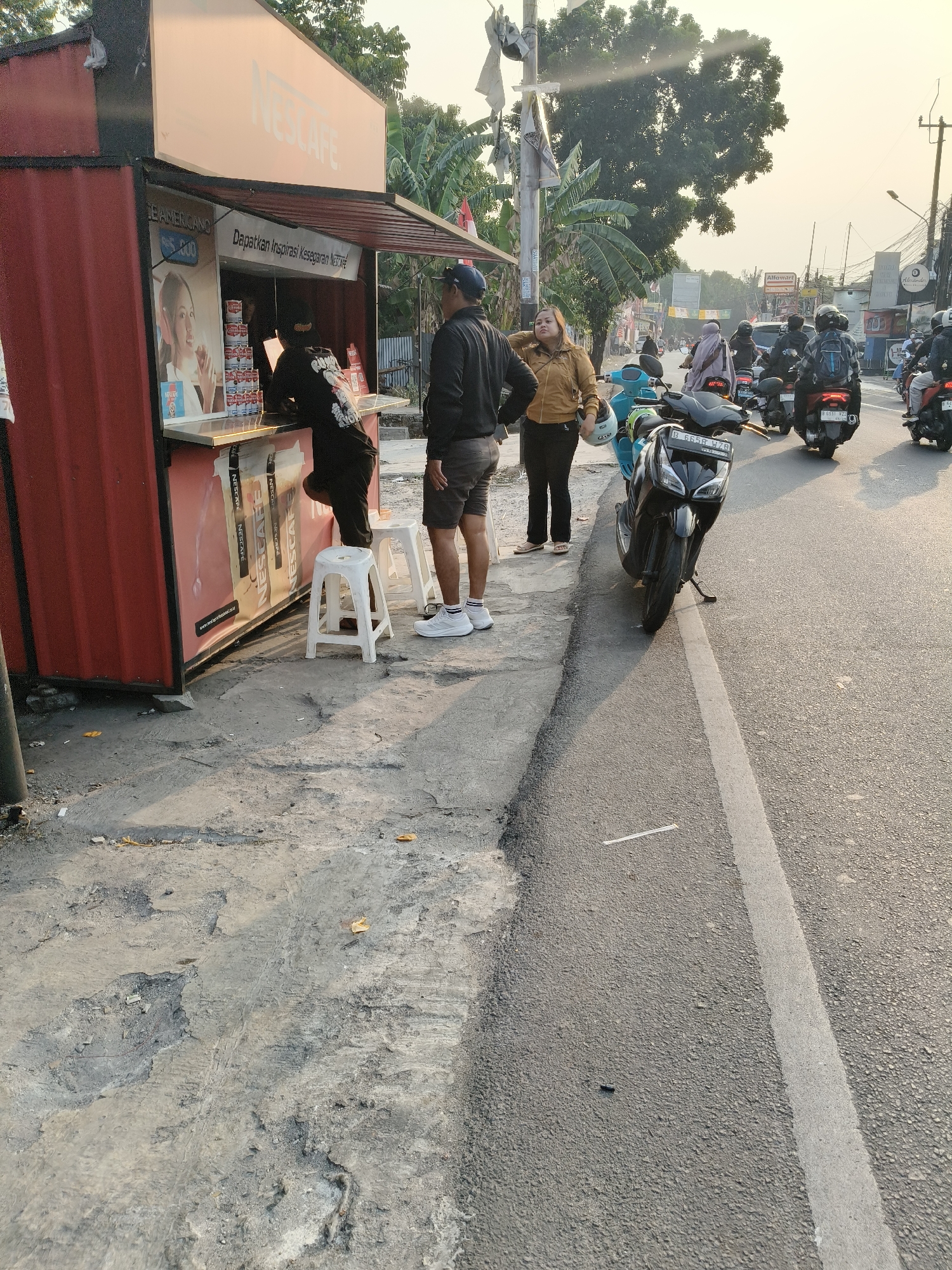 The image depicts a street scene in South Tangerang, Indonesia, at what appears to be dusk or dawn. In the foreground, a red shipping container converted into a Nescafé coffee stand is visible on the left. A woman in a mustard-colored jacket and black pants, holding a white helmet, stands near the stand, looking to the right. Next to her, a man in a black jacket and shorts is also looking right. Inside the stand, a person in a black t-shirt is serving customers. Two white plastic stools are placed in front of the stand. A black scooter is parked on the asphalt road to the right of the stand. In the background, a busy street is filled with motorbikes and a few cars, suggesting moderate traffic. The road has a white line marking. Buildings and trees line the street, and utility poles with wires are also present. The overall environment is a commercial street with a focus on a small food and beverage business.