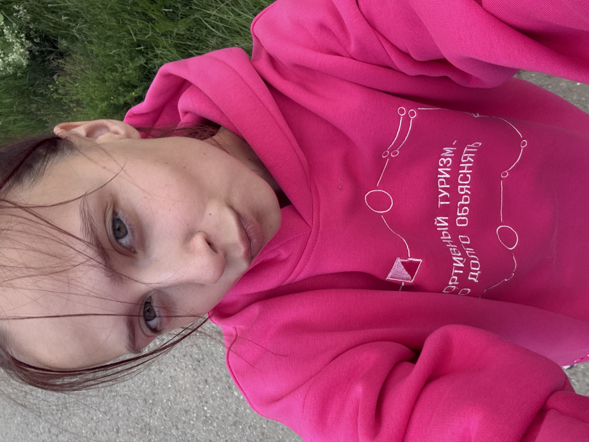 A young woman with light skin and light brown hair tied back, wearing a bright pink hoodie with white embroidered text and a graphic, poses for a selfie outdoors. Her hair is windswept across her face, partially obscuring her eyes. She is looking at the camera with her lips pursed. The background shows a paved surface and lush green grass with some white flowers. The text on her hoodie appears to be in Cyrillic. The general setting suggests an outdoor recreational area or park.