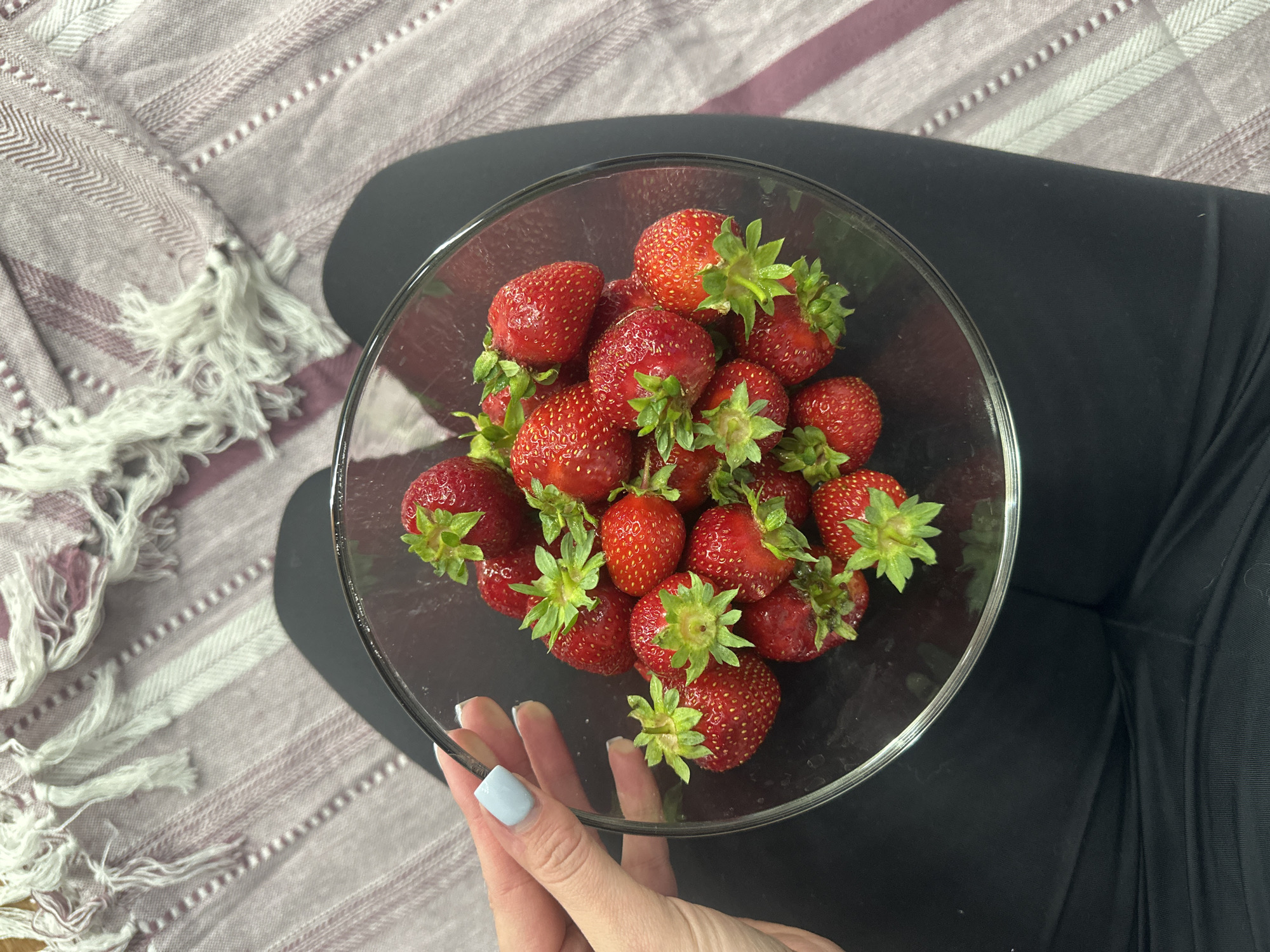 A close-up, overhead shot shows a glass bowl filled with ripe strawberries. The bowl rests on a person's lap, who is wearing black leggings and is sitting on a light purple and white striped blanket. The blanket has white tassels on its left side. The strawberries are a vibrant red with green stems and appear fresh. The scene is brightly lit, suggesting daytime, though the specific time and weather are not discernible. The person's hand, with light blue painted fingernails, is visible at the bottom of the frame, gently holding the edge of the bowl. The setting appears to be indoors or in a shaded outdoor area. No text is visible in the image.