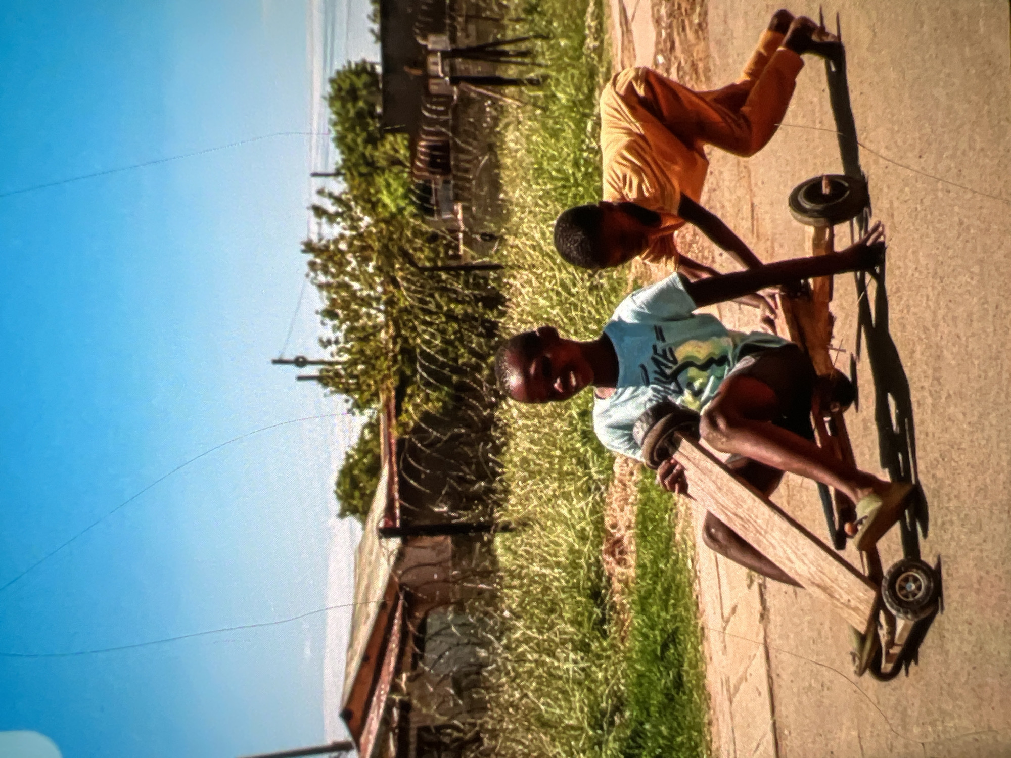 Two young Black boys are playing with a homemade wooden cart on a paved road in a residential area of Lagos, Nigeria. The boy in the foreground, wearing a light blue t-shirt and dark shorts, is seated on the cart, holding onto a wooden steering mechanism. His feet, clad in light green sandals, rest on the front part of the cart. Behind him, another boy in a bright yellow t-shirt and orange pants is pushing the cart, bent over and focused on his task. The cart has four wheels, two visible at the front and one at the rear.
The background depicts a typical Lagos scene. A corrugated iron roofed building, possibly a dwelling, is visible behind a chain-link fence topped with barbed wire. The fence encloses a patch of overgrown, dry grass. Further in the background, more buildings and trees are discernible under a bright, clear blue sky with some faint lines, possibly from the image capture process. The overall environment suggests a sunny day in a part of the city with basic infrastructure. The boys appear engaged in a moment of playful activity, indicative of childhood resourcefulness and enjoyment.