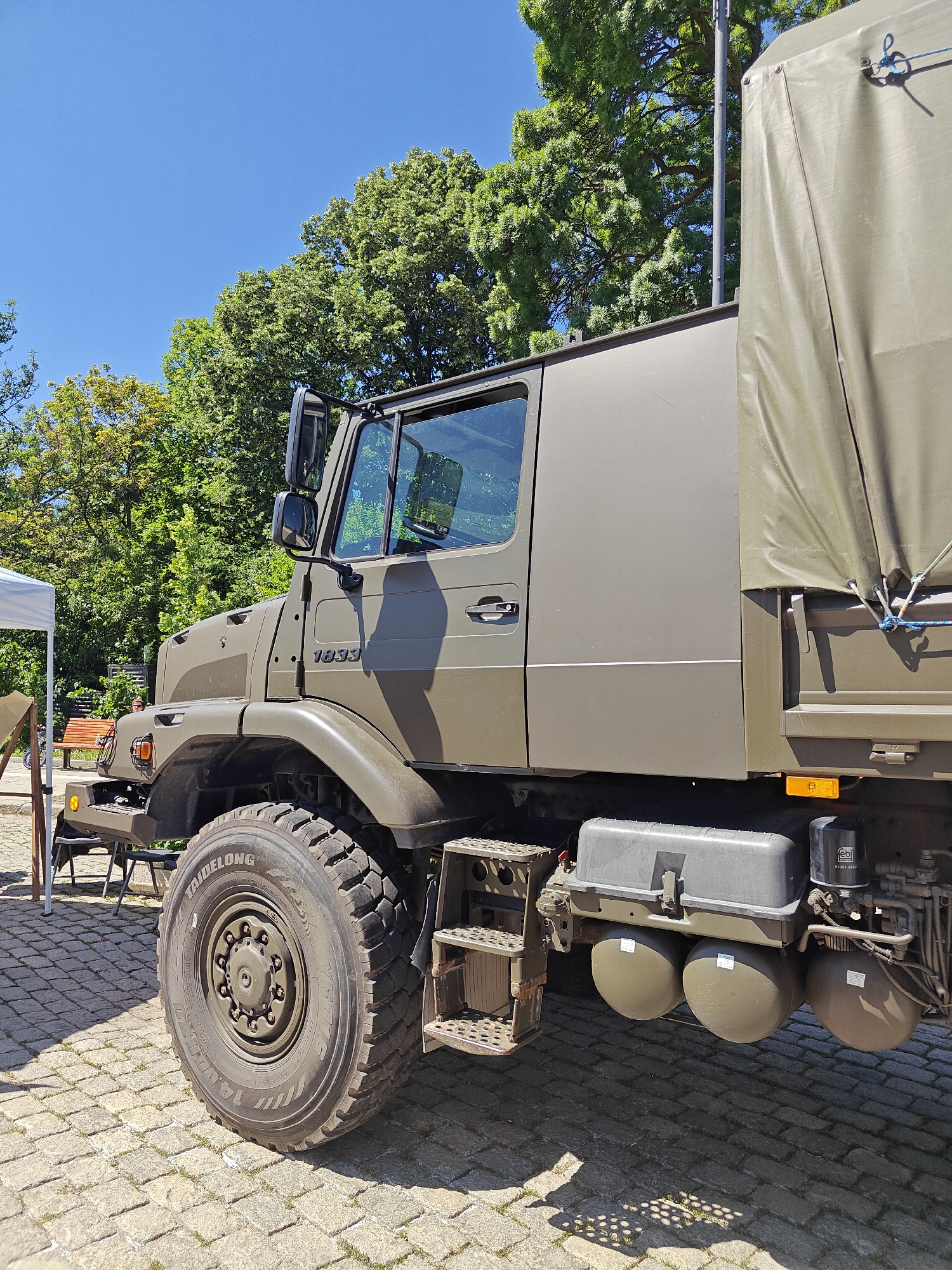 The image shows a military vehicle, specifically a Mercedes-Benz Unimog, parked on a cobblestone surface in what appears to be an urban setting. The vehicle is painted in a matte olive-green color. It has large, rugged tires with the brand name "TAIDELONG" visible. The vehicle is equipped with a passenger access ladder. There is a storage box and several spherical tanks. The trees and blue sky suggest a sunny day. The location is the city of Sofia, Bulgaria.