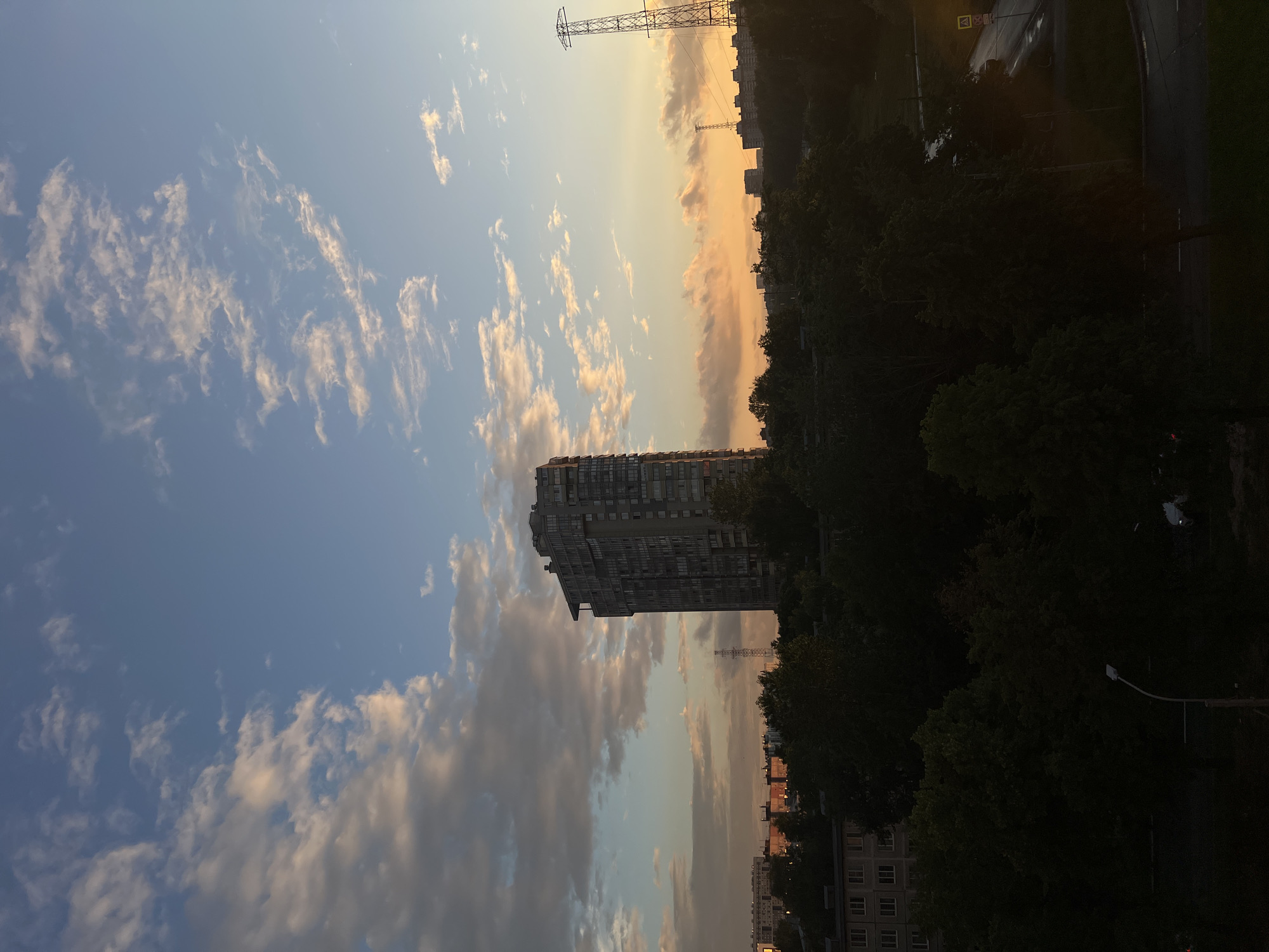 The image shows a cityscape at dusk with a tall residential building in the foreground. The sky is filled with scattered clouds, some illuminated by the setting sun with hues of orange and yellow. In the background, more buildings and a power line tower are visible, silhouetted against the sky. Trees line the lower part of the image, casting shadows. The scene suggests an urban environment, likely a residential area in Saint Petersburg, Russia, during the evening. There are no visible people or specific activities taking place, but the warm colors in the sky evoke a sense of calm and the end of the day.