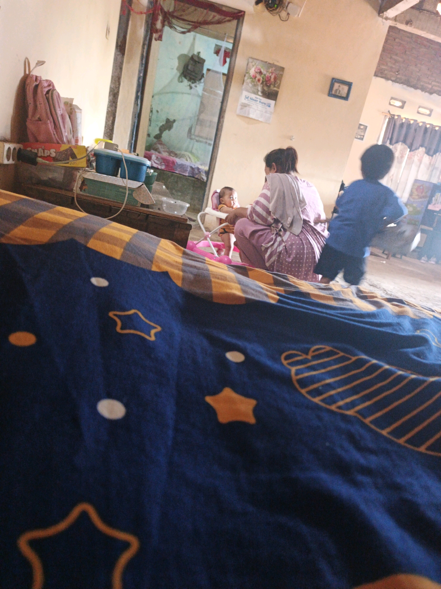 The image depicts an indoor scene that is likely a home in Bumiharjo, Indonesia. In the foreground, a blue and yellow striped blanket is spread out, partially obscuring the view of the rest of the room. In the mid-ground, a woman in a purple and white patterned dress is holding a baby in a pink baby bouncer. Behind them, a young child in a blue shirt and dark shorts is walking away from the camera. The room appears to be sparsely furnished, with a table and chairs visible in the background. The walls are painted a light yellow, and the ceiling is made of wood. The overall impression is one of a simple, domestic setting.