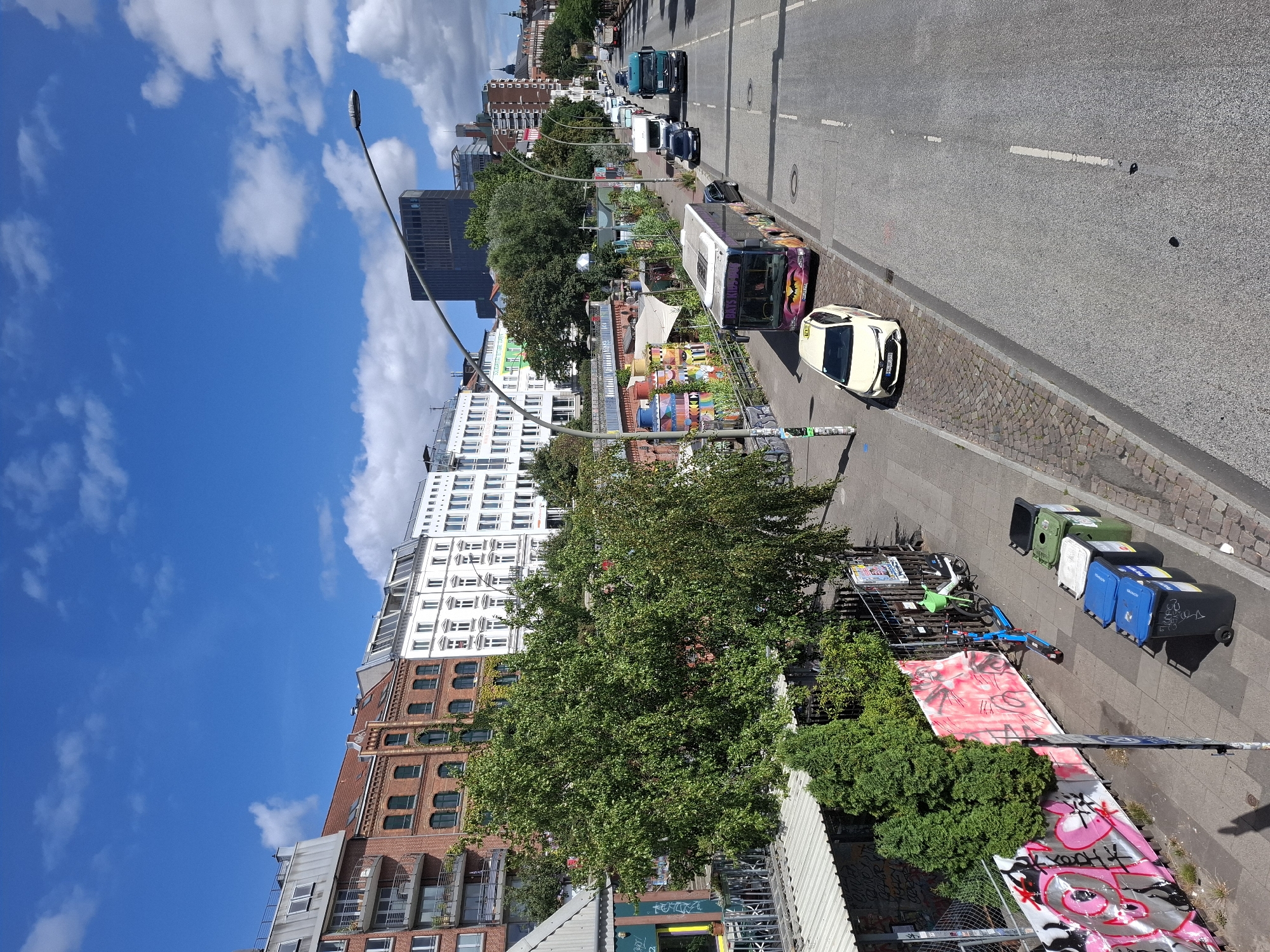 The image depicts a street scene in Hamburg, Germany, likely along the Elbe riverfront or a similar urban area. The perspective is from an elevated position, looking down on a street with several vehicles and buildings.
On the left side of the street, there are several multi-story buildings with a mix of architectural styles, including red brick and white facades. Some buildings have balconies and numerous windows. In front of these buildings, there is a significant amount of greenery, including large trees and smaller bushes. A fence with graffiti-covered panels separates the sidewalk from the buildings. Along the sidewalk, there are several garbage bins, a blue electric scooter, and a white bicycle with a green basket.
The street itself is paved with asphalt and has white lane markings. Several vehicles are visible on the road, including a yellow taxi, a purple bus with colorful graphics, a dark-colored car, a white camper van, and a large green truck. The road appears to be a main thoroughfare, with traffic moving in the distance.
In the background, more buildings can be seen, including a modern, dark-colored skyscraper. The sky is bright blue with scattered white clouds, indicating a sunny day. The overall impression is a lively urban environment with a blend of residential, commercial, and recreational elements.