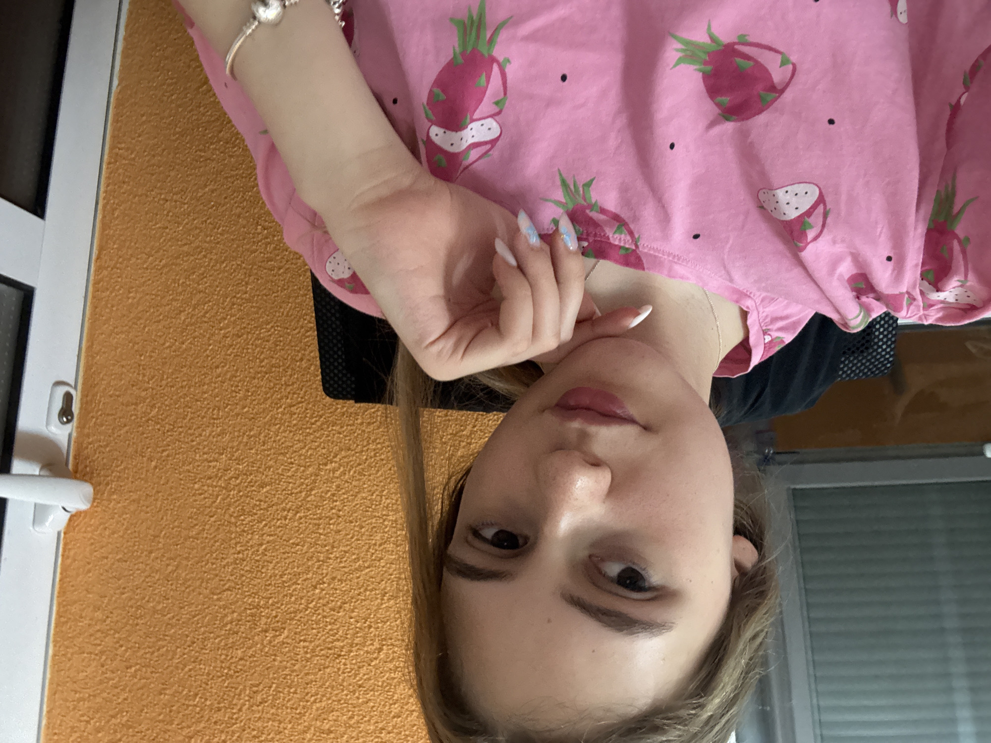 A young woman is centered in the image. She is facing the camera with her hand under her chin. She is wearing a pink, dragon-fruit patterned shirt. She has fair skin, brown eyes, and long brown hair. She is wearing a bracelet on her left wrist. Behind her is a wall with orange stucco and a closed window. The city is not visible from this picture. Panagyurishte, Bulgaria cannot be verified from this image.