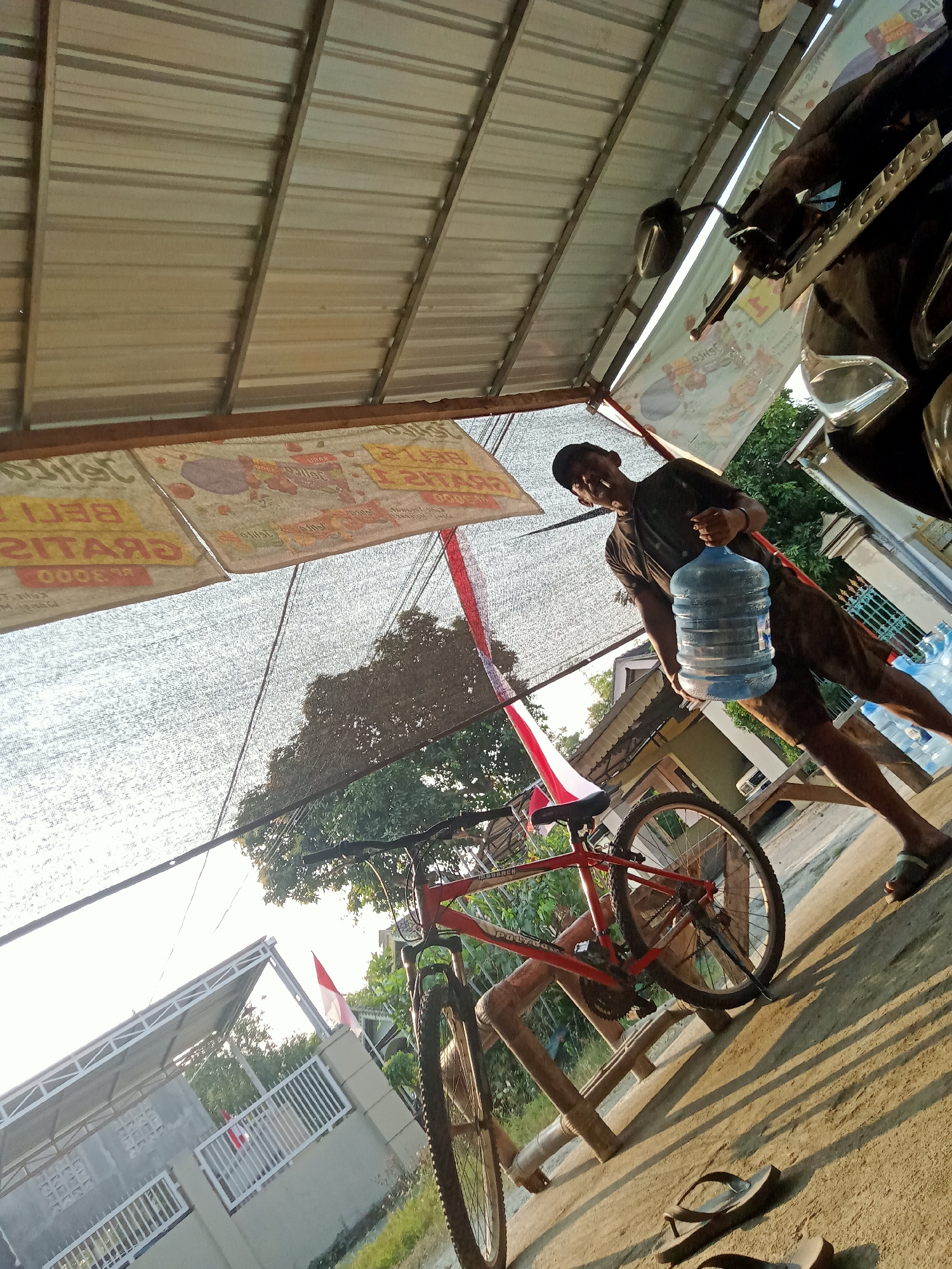 A man wearing a dark t-shirt, dark shorts, and a dark baseball cap is standing outdoors, holding a large blue water jug in his right hand. He is in what appears to be a covered outdoor area, possibly a marketplace or a roadside stall, with a corrugated metal roof overhead. Behind him, a red bicycle is propped up on a wooden stand. Several more water jugs are visible in the background, along with a motorcycle parked to the right. A banner with text in Indonesian is hanging overhead. The scene is illuminated by the warm light of what seems to be late afternoon or early morning sun, casting long shadows on the ground. In the distance, houses and trees are visible, and Indonesian flags are displayed on a building. The setting suggests a humid, tropical environment typical of Simo, Indonesia.