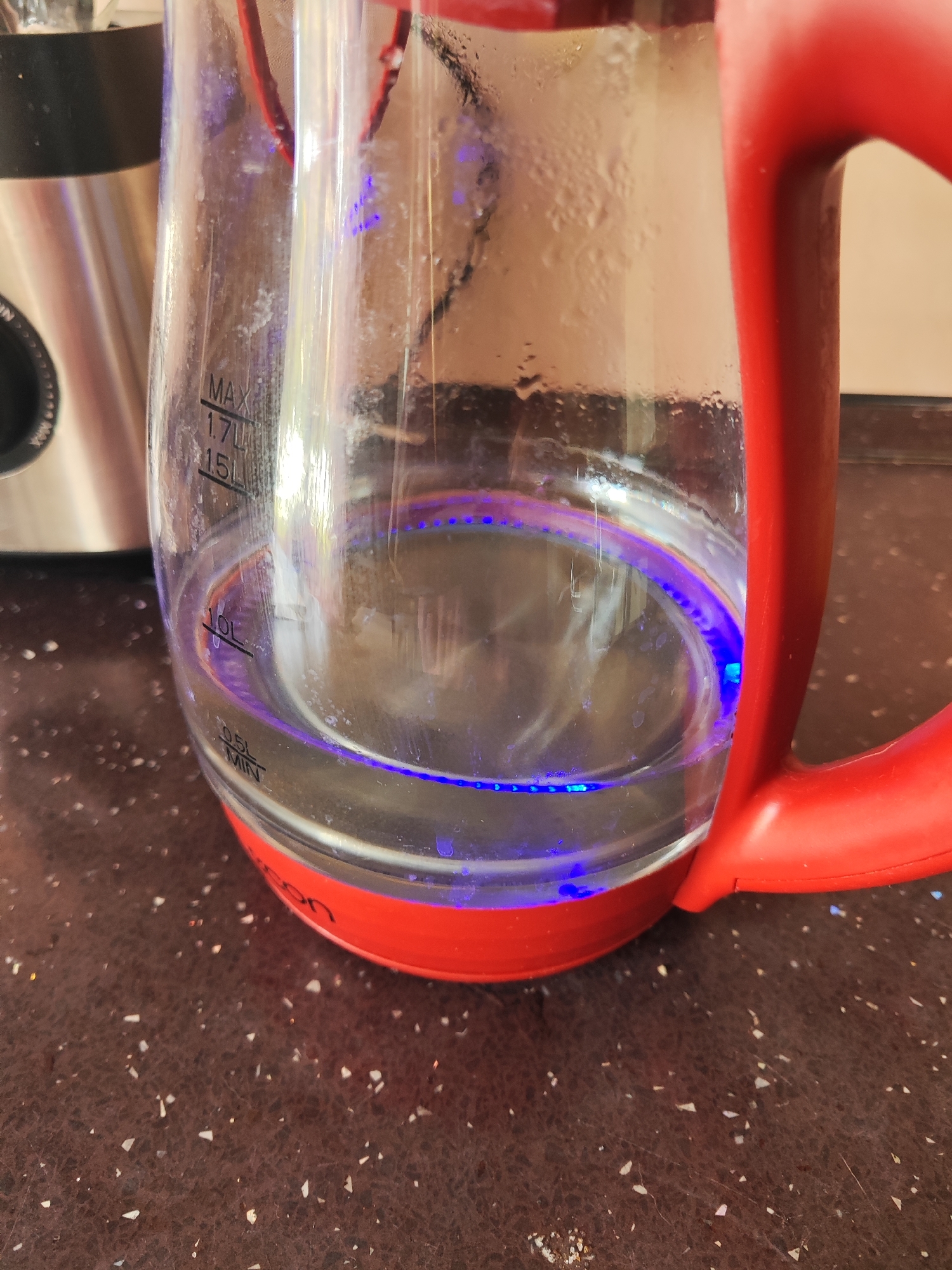 An interior shot, possibly in a kitchen, captures a clear glass electric kettle with a bright red handle, sitting on a dark speckled countertop. The kettle contains water, with a blue light emanating from the base, indicating it is likely in operation or has recently been used. Measurements are visible on the glass: MAX 1.7L, 1.5L, 1.0L, and MIN 0.5L. Condensation is present on the upper interior of the kettle. The background is blurred, but a metallic appliance is partially visible to the left. There is no indication of any specific city or location within the image.