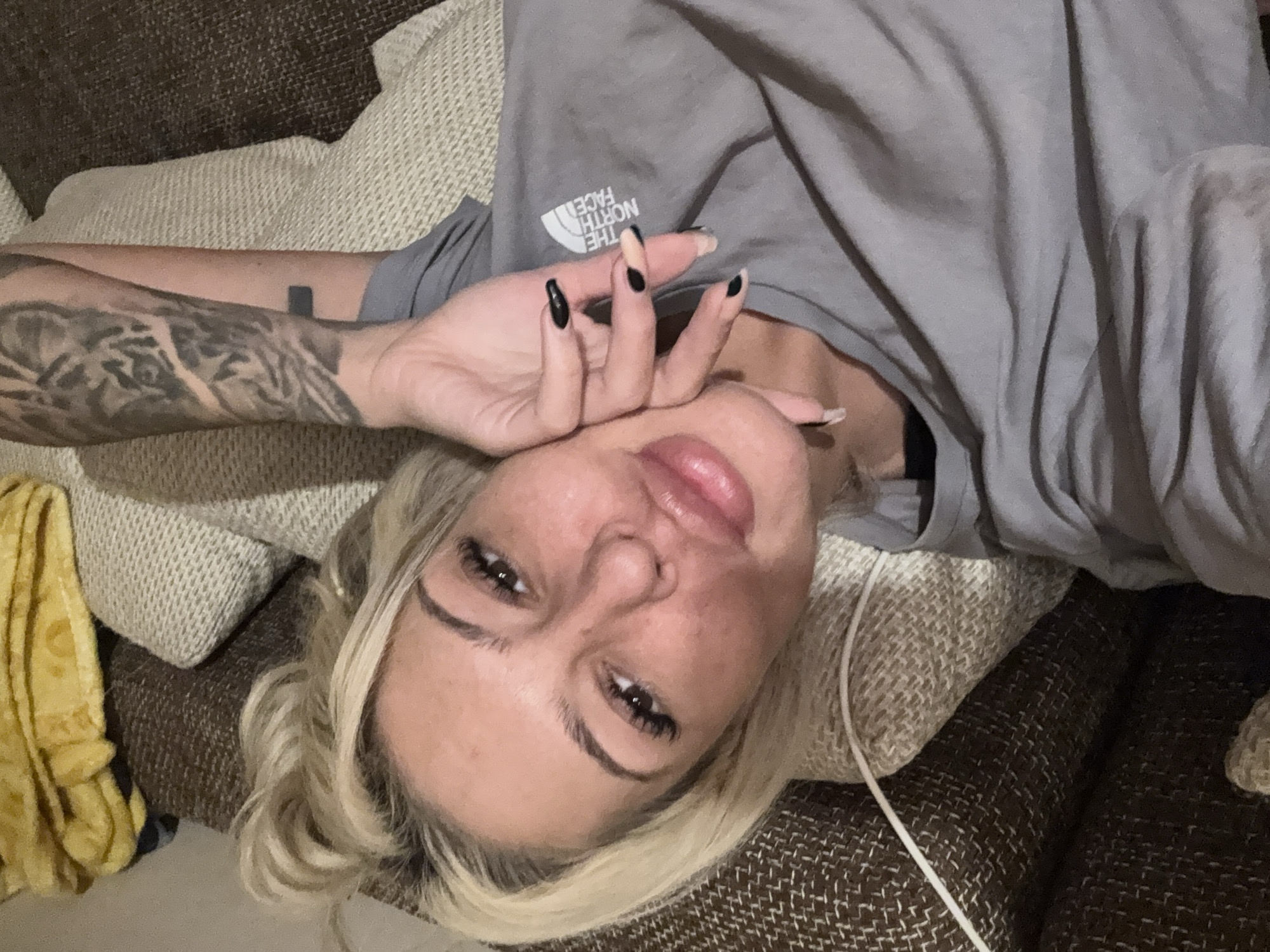 An indoor, high-angle close-up captures a blonde woman lying on a sofa, looking directly at the camera. The setting appears to be a comfortable lounge area, possibly in Plovdiv, Bulgaria, given the textured brown and beige upholstery of the couch. She is wearing a grey t-shirt with the upside-down white logo "THE NORTH FACE" visible on the chest.
Her right arm, which is heavily tattooed with a dark, intricate design, is raised, with her hand gently touching her face near her mouth and chin. Her fingernails are long and pointed, with most painted black, but at least one on her right hand is a contrasting nude or light beige color. Her lips are slightly parted, and she has visible eyeliner and mascara. Her expression is calm and neutral, possibly a slight smile. A yellow blanket or textile is partially visible to the left of her head. A thin white cable or string runs across the sofa near her chest. The lighting suggests an indoor evening or nighttime setting.