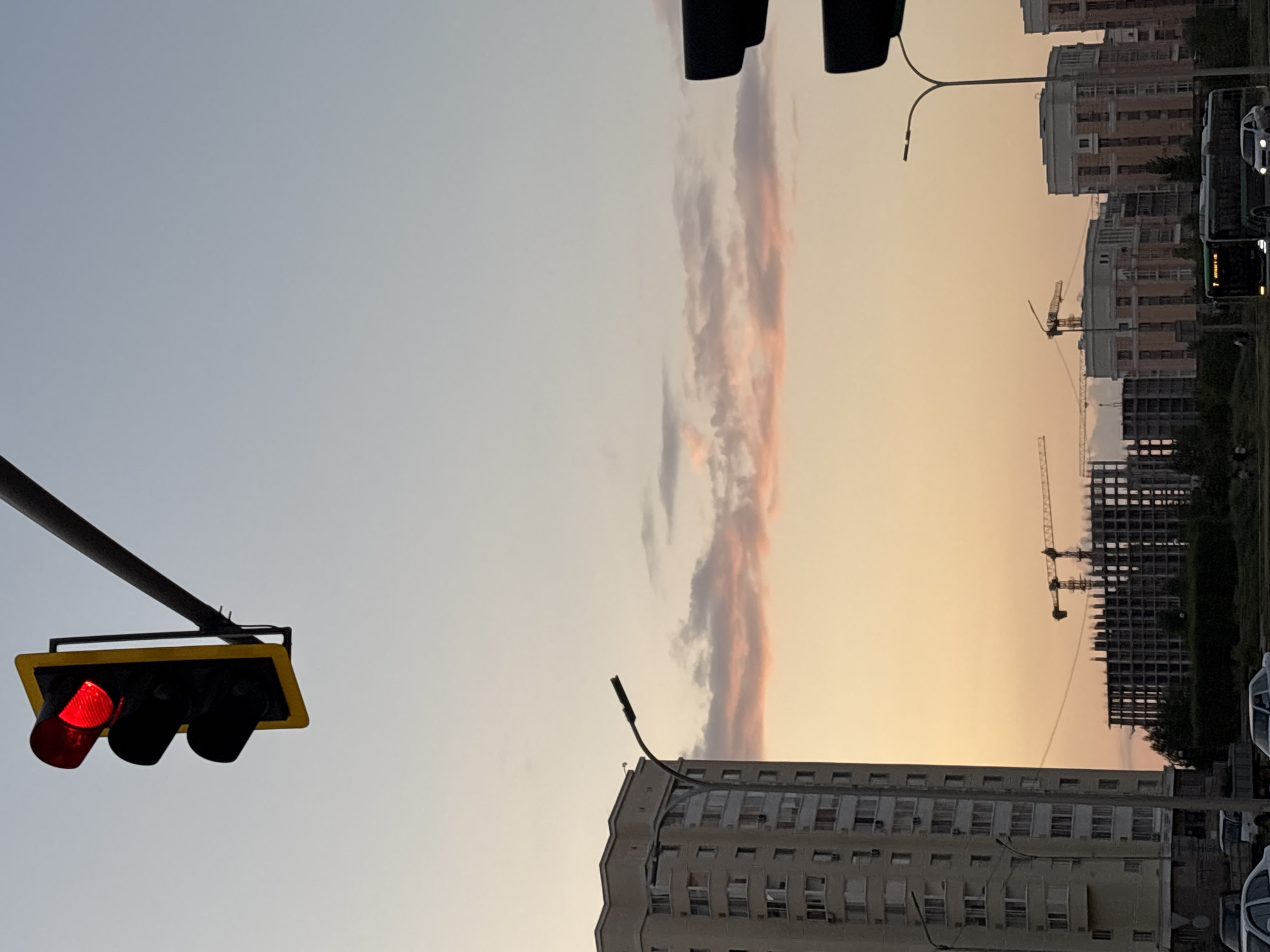 The image depicts a city scene, likely in Astana, Kazakhstan, at dusk. A traffic light with a red signal is prominently displayed in the upper left quadrant, suspended from a metal pole. In the middle ground, a tall, light-colored apartment building with numerous windows stands to the left. Further into the background, multiple construction sites are visible, with unfinished buildings and cranes. A street lamp curves over a road in front of the apartment building. The sky exhibits a gradient of soft, warm colors, with pinkish clouds against a pale blue expanse, indicating sunset. In the foreground, vehicles, including a bus and cars, are present on a road. The overall atmosphere is urban, with elements of daily life and ongoing development.