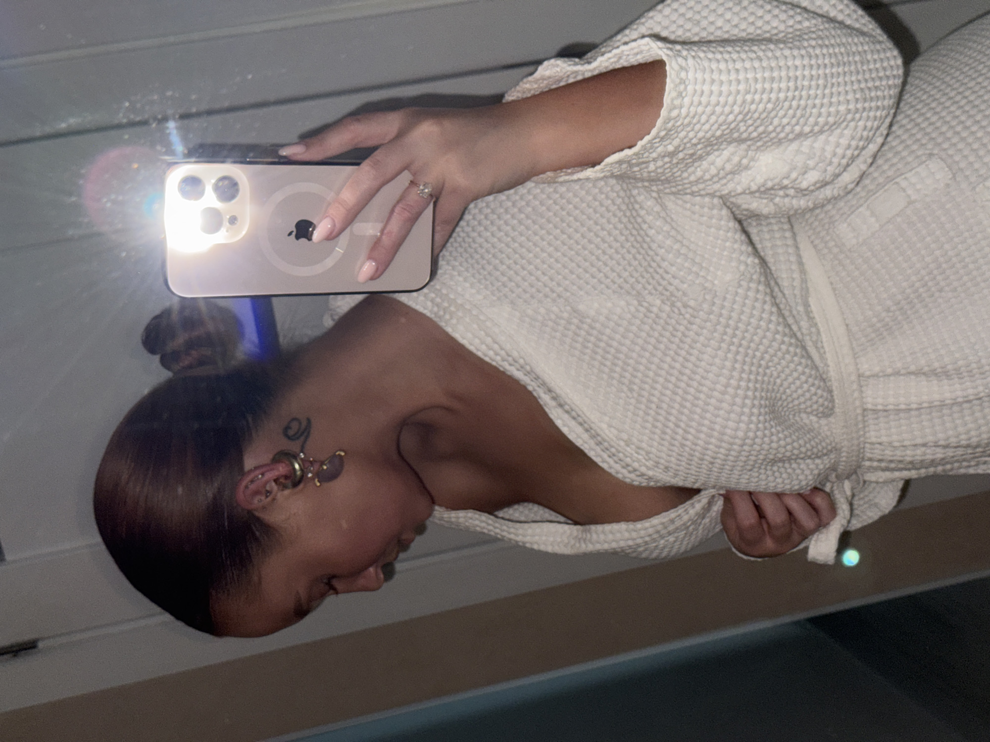 A woman in a white waffle-weave bathrobe is taking a mirror selfie with an iPhone, the flash of which is reflecting in the mirror. She has brown hair pulled back in a bun, and a tattoo is visible near her ear. She is wearing a ring on her finger. The background is partially visible, with light-colored horizontal panels at the top and darker, indistinct surfaces below. There is no information about the location in the city of Chaikovsky, Russia.