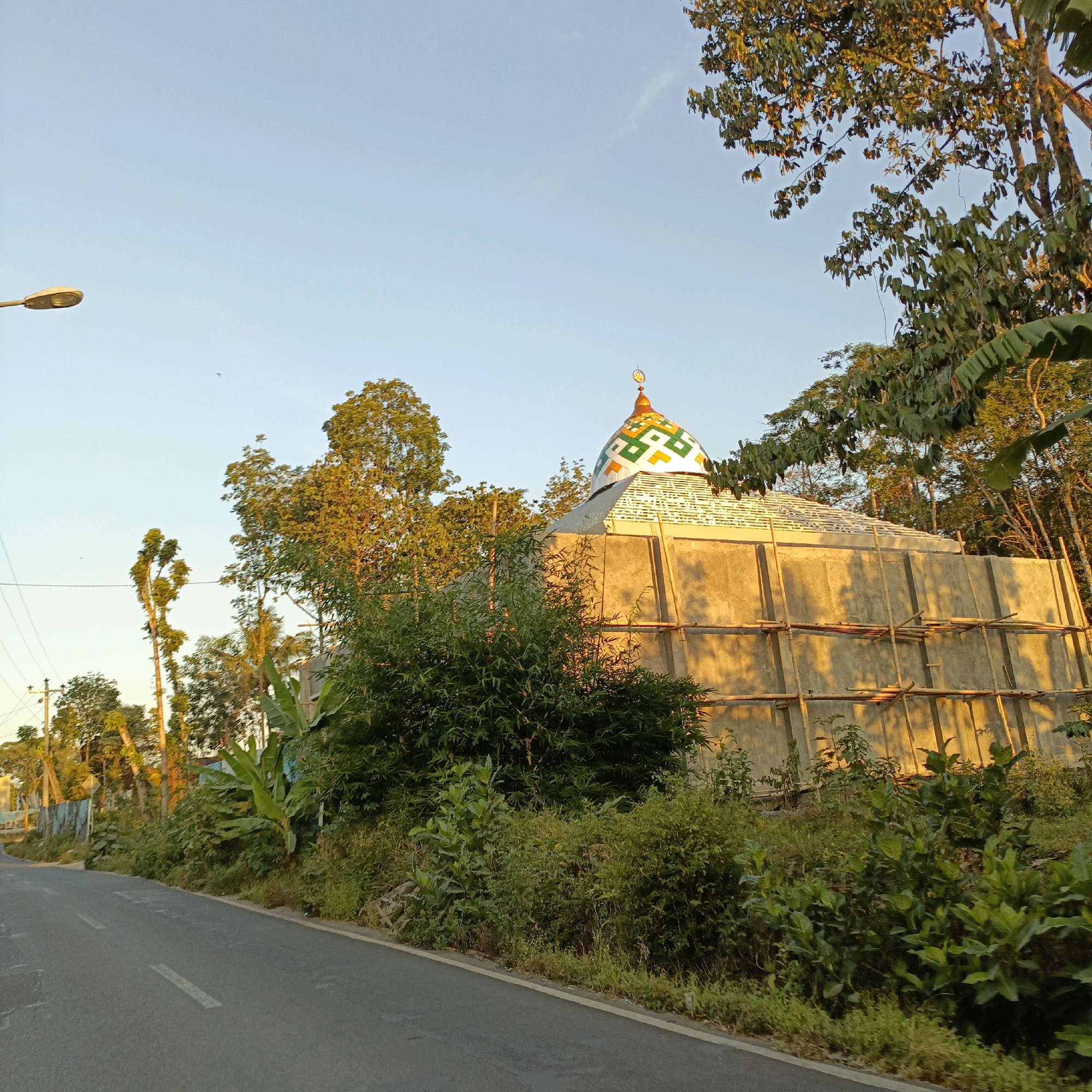 This is a full-shot image of a mosque under construction, located on the side of a road in Salatiga, Indonesia. The mosque has a distinctive dome with a colorful pattern of white, green, and yellow geometric shapes, topped with a crescent moon. The building itself is made of concrete, with scaffolding made of bamboo poles surrounding its exterior walls, suggesting ongoing construction work.
The environment around the mosque is lush and green, with various types of trees and dense vegetation growing along the roadside and behind the building. In the foreground, a paved road with white lane markings stretches into the distance, curving slightly to the left. A street lamp is visible on the left side of the road, extending over the shoulder. The sky is a clear, pale blue, indicating it's either early morning or late afternoon, given the warm, golden light illuminating the side of the mosque and the surrounding foliage. No people are visible in the image.