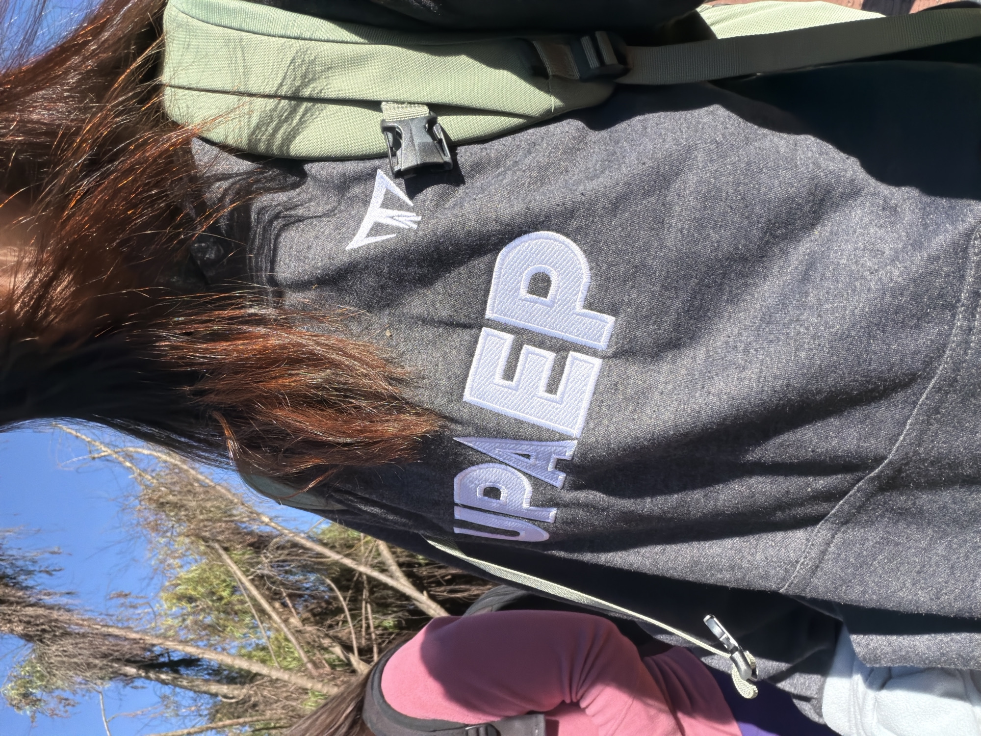 The image shows a close-up view of a person wearing a dark grey hooded sweatshirt with white lettering that reads "UPAE P". The person is also wearing a green backpack. In the background, there are trees and a blue sky. Another person wearing a pink sweatshirt is partially visible on the left. The scene appears to be outdoors. The location is the municipality of Bojacá, Cundinamarca, Colombia.