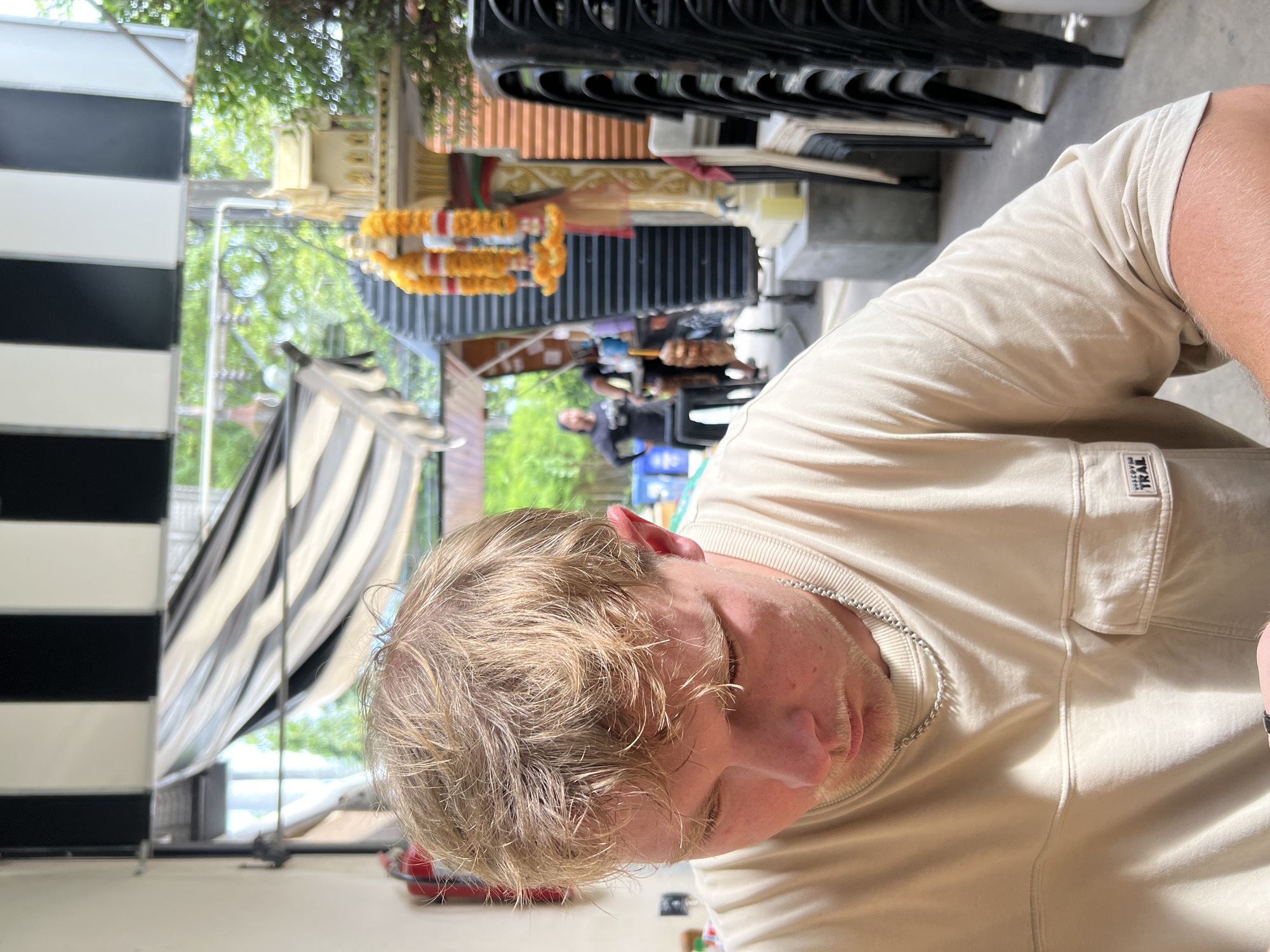 The image depicts a young Caucasian man with light brown hair and stubble, wearing a light tan crewneck t-shirt and a silver chain necklace. He is looking down and to his left. In the background, an outdoor seating area is visible with stacked black chairs. A shrine adorned with orange and white garlands is also present, along with a striped awning overhead. Two individuals, appearing to be of Asian descent and wearing dark clothing, are in the mid-ground. The setting suggests an establishment in Choeng Thale, Thailand, likely a restaurant or market, given the outdoor ambiance and Thai cultural elements.
