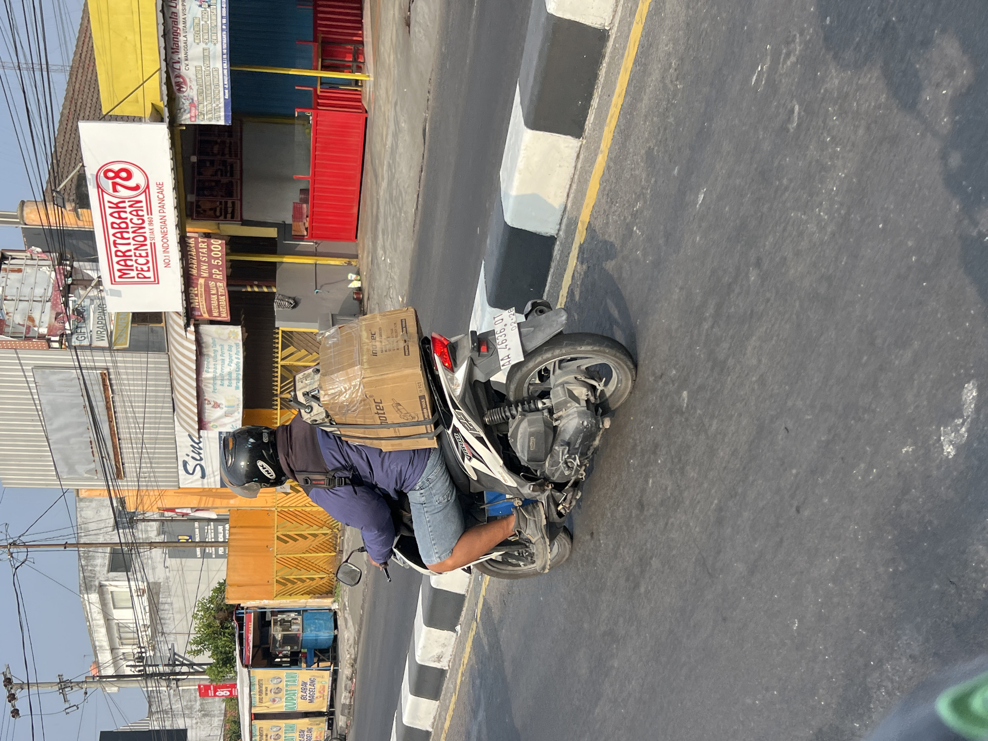 A person wearing a dark blue jacket, denim shorts, and a black helmet rides a white and black motorcycle on a paved road. The motorcycle has a large cardboard box strapped to its rear, secured with clear plastic wrap. The box has text and images on it, including "InnoTec" and what appears to be a picture of a power tool. The person's right leg is bent, with their foot resting on the motorcycle's footrest. The motorcycle's license plate is visible and reads "AA 4636 QT". The road is bordered by a curb painted with alternating black and white sections. In the background, buildings line the street, including a building with a yellow awning and a sign that says "MARTABAK PECENONGAN 78". Another sign reads "CV. MANGGALA UTAMA VISIPRIMA". There are also food stalls and power lines visible. The sky is clear and blue, suggesting it is daytime. The scene is located in Sinduadi, Indonesia, as indicated by the text on the sign.