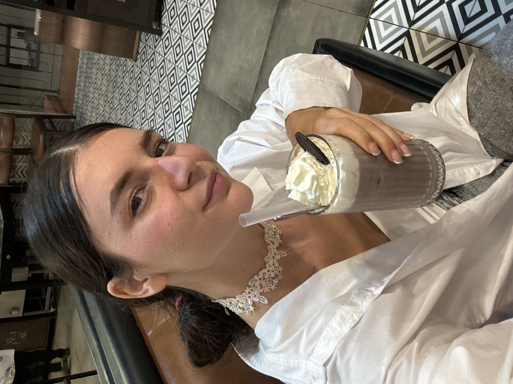 A young woman with dark hair pulled back and a pink tie is holding a chocolate drink with whipped cream and a cookie. She is wearing a white button-down shirt and a lace choker with pink and white flowers. She is seated in a dark leather chair, in what appears to be a cafe or restaurant. The flooring is a geometric pattern of black, white, and grey tiles. In the background, there are tables and chairs, suggesting an indoor dining area. The lighting is warm and ambient. The location is identified as Phan Thiết, Vietnam.
