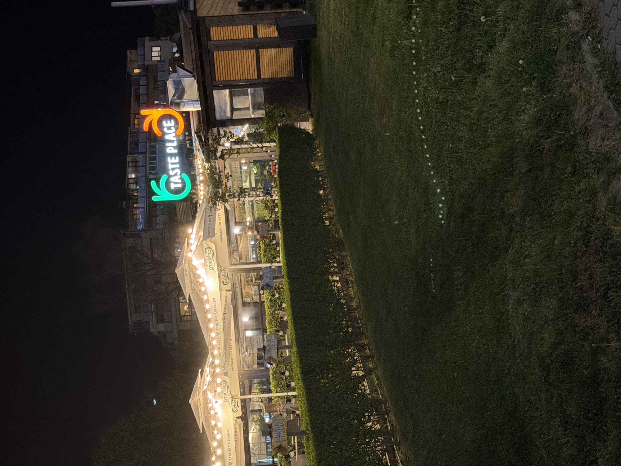 The image presents an outdoor scene at night, apparently taken at a restaurant called "Taste Place," recognizable by its prominent neon sign featuring the name and stylized "taste" symbols. The restaurant has an outdoor seating area with multiple white canopies, likely covering tables for dining, and string lights are visible, contributing to the ambiance. A hedge borders the seating area, separating it from a grassy lawn, which is also illuminated with a row of small lights. In the background, the silhouettes of buildings can be seen. The image could possibly be located in the city of Sofia, Bulgaria, as the Cyrillic alphabet is visible on the awnings.