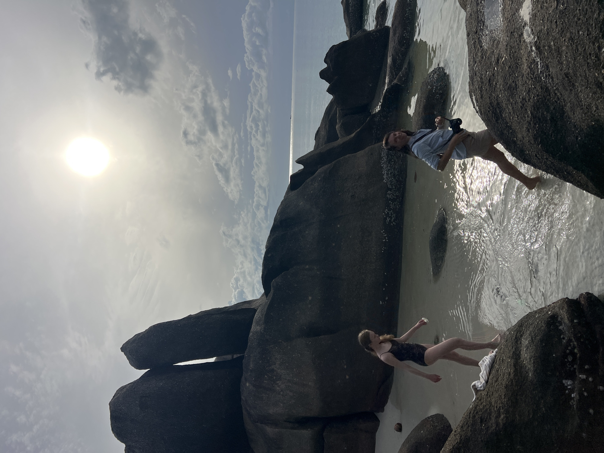 The image depicts two individuals on a rocky beach in Baan Chaweng Noi, Thailand. In the foreground, a person with long brown hair, wearing a light blue shirt and tan shorts, is walking towards the viewer with a camera around their neck. To the left and slightly behind, a person with long blonde hair, wearing a dark patterned one-piece swimsuit, is walking away from the viewer. Both are wading in shallow water among large, dark, rounded rocks. The background features a bright sun in a cloudy sky, the ocean, and more large rock formations. The tide appears to be low, exposing the wet sand and rocks.