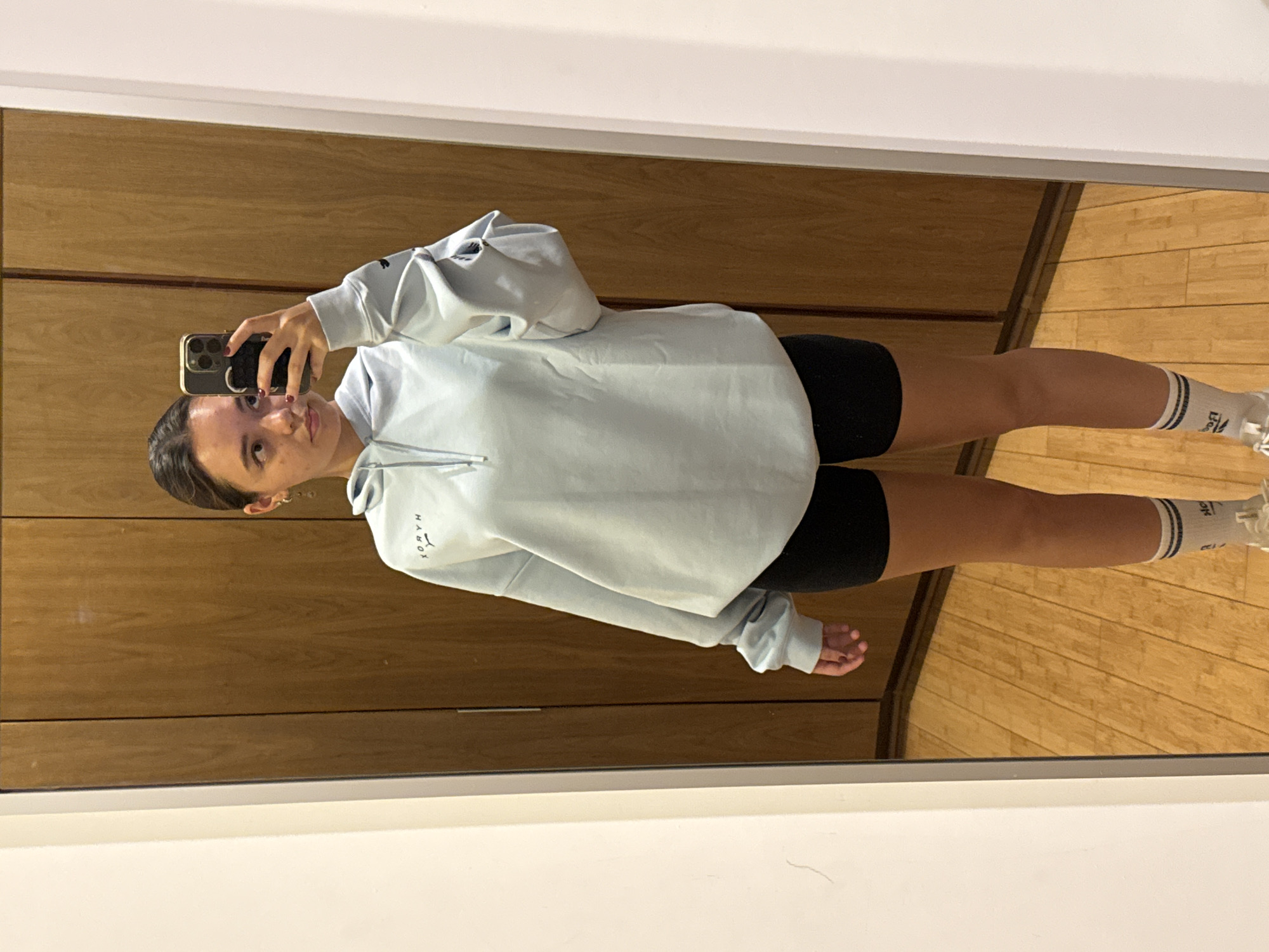 A person is visible in a mirror selfie. The person is wearing a light blue hoodie and black shorts with white socks and sneakers. The background shows wooden cabinets and a wooden floor. The location is not San Bernardino Tlaxcalancingo, Mexico.