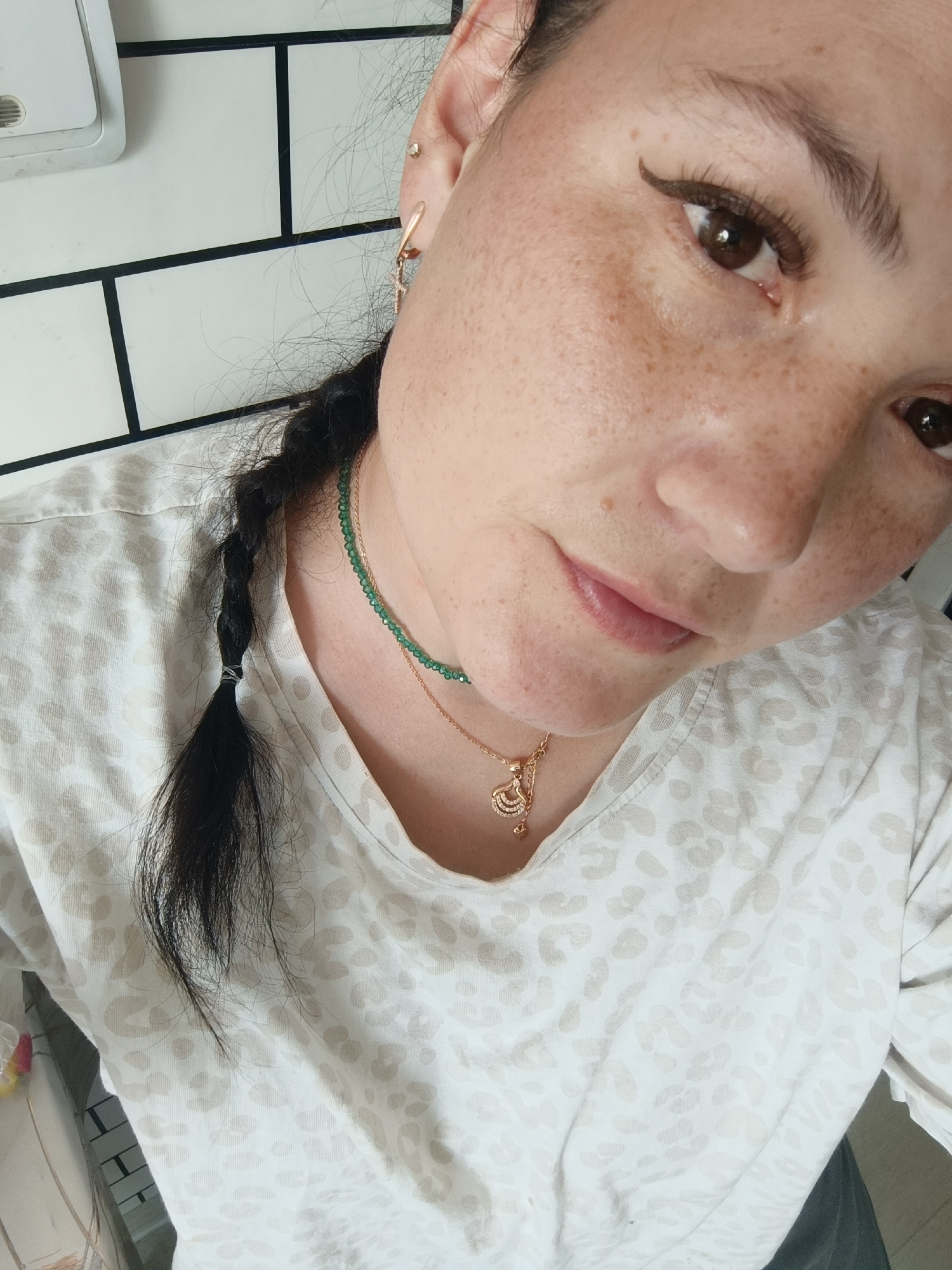 A close-up, waist-up shot shows a person with fair skin and dark hair, which is styled into a braid. They are wearing a white shirt with a light brown leopard print pattern and two necklaces. One necklace is made of small green beads, and the other is a delicate gold chain with a small pendant shaped like a fan. The person has a small stud piercing in their upper ear and a dangling earring in their lower ear. They are also wearing eyeliner. The background appears to be a tiled wall with dark grout lines, possibly a bathroom or kitchen. The lighting is natural, casting soft shadows on the person's face and neck. There is no specific indication of the location being in Chaikovsky, Russia.