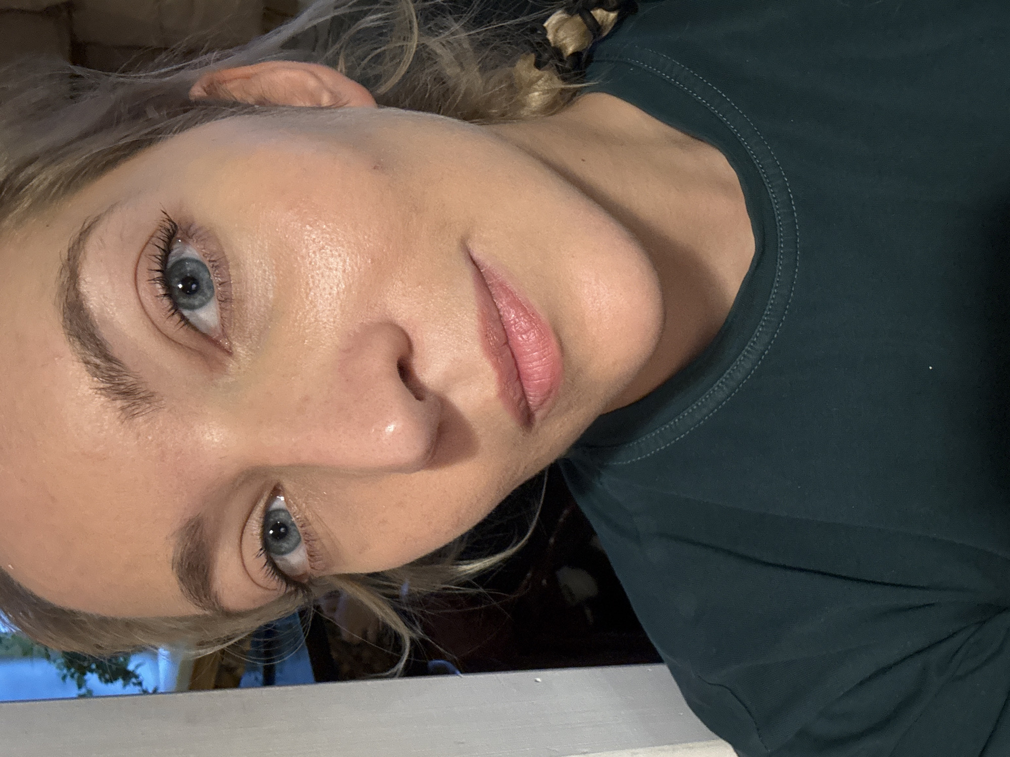 A woman is pictured in a close-up, frontal view, with the camera positioned slightly below eye level. She has blonde hair styled in a braid that is visible on her right side. Her skin is light-toned with a dewy finish. Her eyes are blue and appear to be looking slightly to the left of the camera. She is wearing a dark green t-shirt. The background is dark and out of focus, with a vertical white object on the left side of the frame that could be a door frame or wall. There is a hint of greenery visible through a window behind her on the left. The image appears to have been taken indoors.