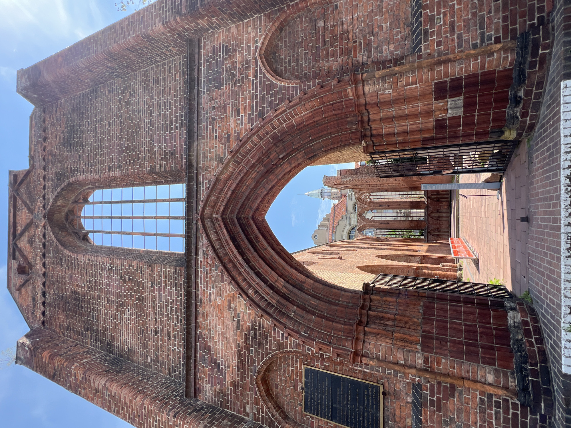 The image shows the ruins of the St. Nikolai Church in Berlin, Germany. The church is made of red brick and has a large arched entrance. Through the arch, more of the ruined church can be seen, including tall windows and other brick structures. A tall, slender spire is visible in the distance, and other buildings with red tiled roofs are also present. The ground in front of the church is paved with red bricks. There are black metal gates on either side of the entrance, and a black plaque is mounted on the left wall. A white railing is visible in the foreground. The sky is blue with some clouds. There are no people visible in the image.