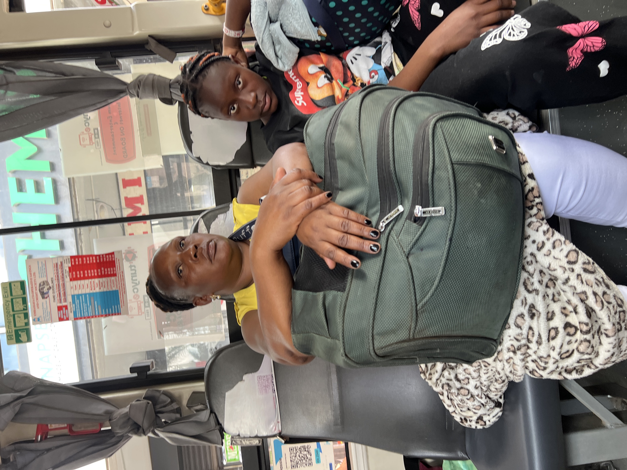 The image depicts two African women and a large backpack inside a bus. The bus is presumably within the city of Nairobi, Kenya, based on the "Nairobi" and "NAPSA" seen on an advertising sign within the bus. The older woman, wearing a yellow shirt and dark braids, is seated and clutching a large, olive-green backpack. The younger woman, a child with braids and wearing a black t-shirt with a red and white design, is also on the bus, seated to the older woman's left. Both women are looking towards the camera. The interior of the bus is visible, including seats, windows with advertisements, and curtained overhead storage. A leopard-print blanket is visible between the legs of both women.