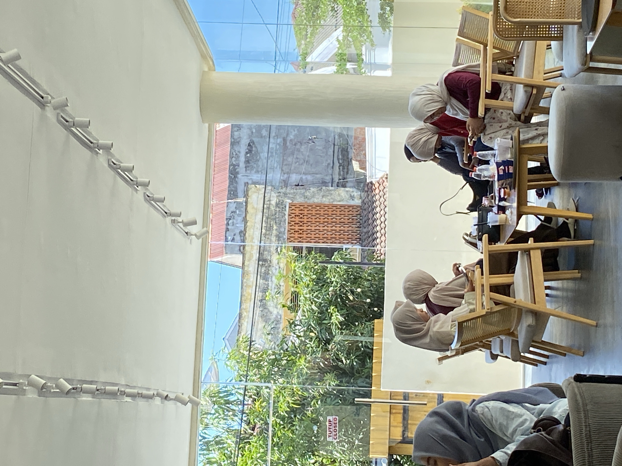 The image shows an indoor scene, possibly a cafe or restaurant, in Lhokseumawe, Indonesia. There are several women wearing hijabs seated at wooden tables and chairs. Some are looking down at objects on the table, possibly interacting with phones or food. There is a large window with a view of the outside, including trees and buildings. The building exterior visible outside the window appears to be residential. The interior features white walls with rows of track lighting.