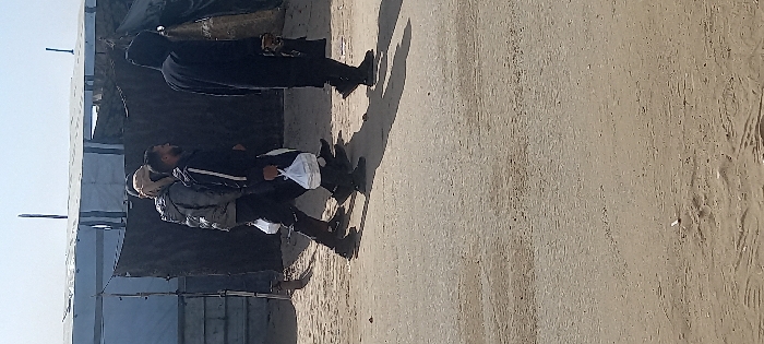 The image depicts three adult males walking from right to left on a dirt path in Khan Yunis, Palestinian Territory. The ground surface is uneven, light brown compacted earth with visible traces of movement and small debris.
From left to right:
1. A male subject, wearing a grey quilted jacket, dark trousers, and a light-colored head covering, carries a white plastic bag in his left hand. His face is partially visible.
2. A male subject, with a dark beard, wears a black tracksuit featuring white stripes on the outer leg seams and dark athletic shoes. He also carries a white plastic bag in his left hand. His profile is visible.
3. A male subject, clad in a long, dark hooded garment that conceals his face, carries an obscured dark object in his right hand.
All three individuals cast distinct shadows onto the path, suggesting a light source from their right. In the background, a row of improvised structures, consistent with temporary shelters or tents, is visible. These structures appear constructed from light-colored tarpaulins or corrugated sheets for roofs and walls, complemented by darker fabric or tarpaulin sections for additional walls. Vertical poles support these structures under a clear, light blue sky.