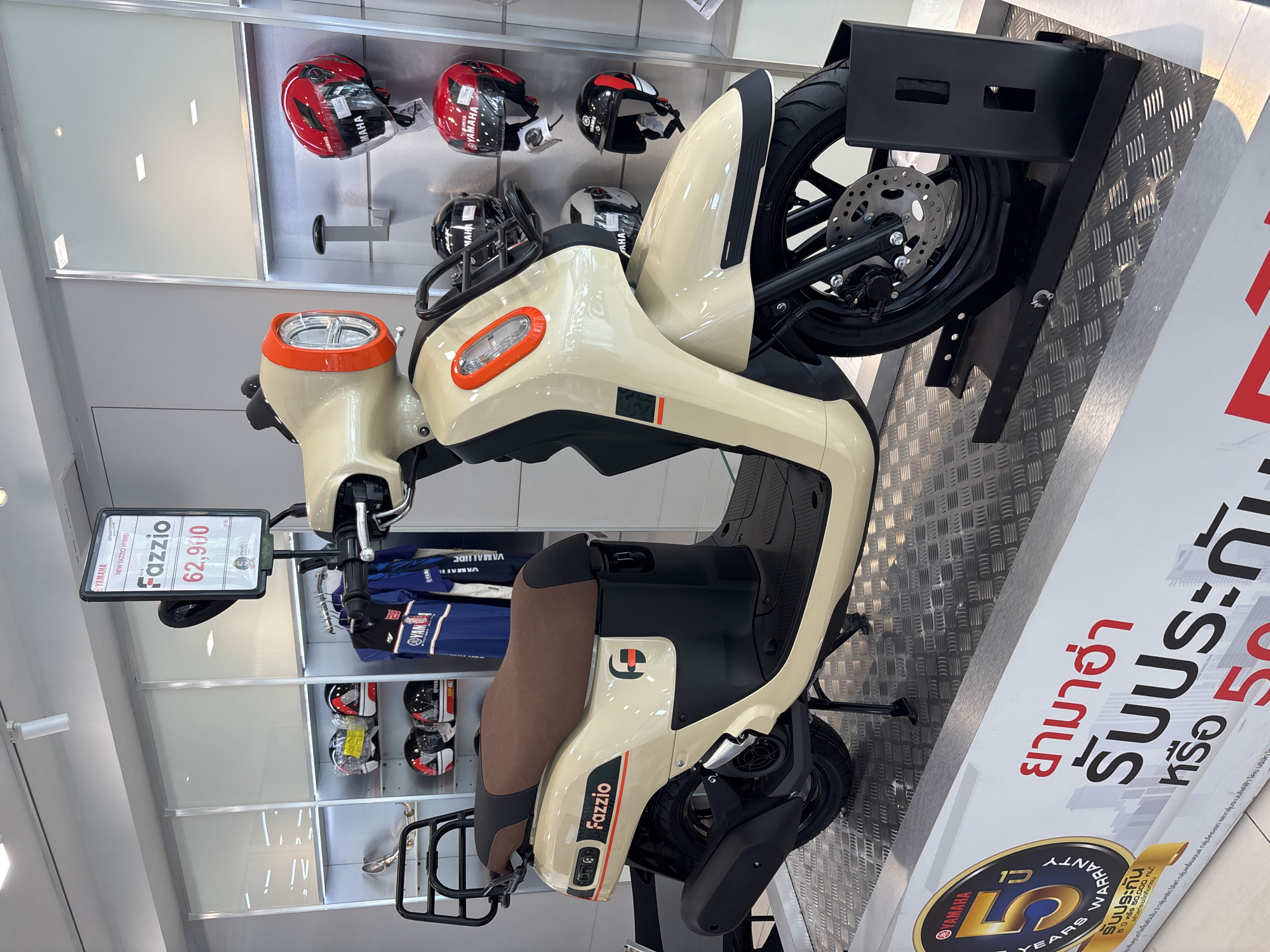 This is an indoor image of a Yamaha Fazzio scooter on display in a showroom. The scooter is beige with orange accents and a brown seat. Behind the scooter are shelves with various helmets and Yamaha apparel. A sign next to the scooter indicates a price of 62,900 and promotes a 5-year warranty. Text on the sign is in Thai. The location is likely a Yamaha dealership in Baan Bo Phut, Thailand.
