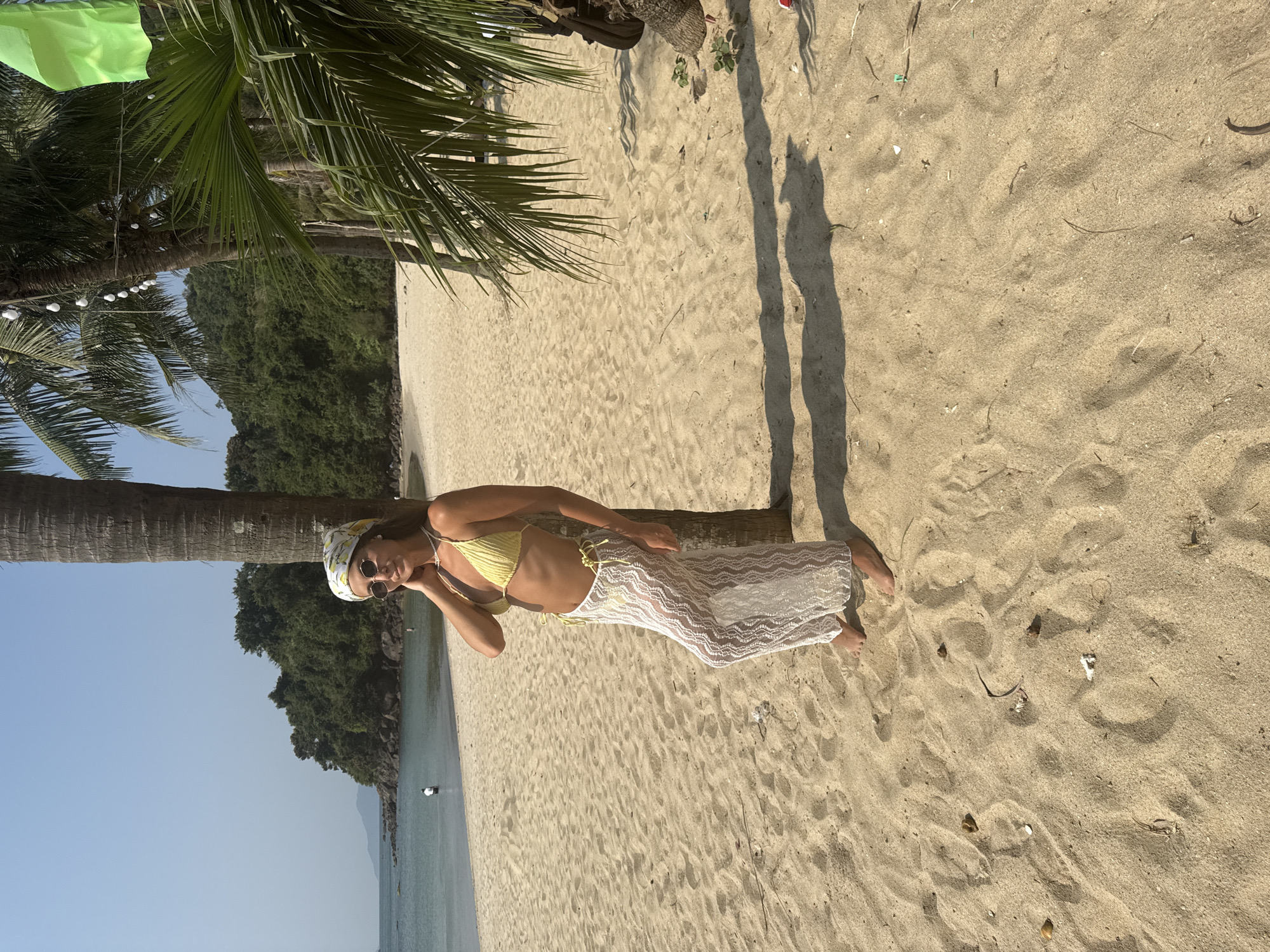 A woman is posed on a sandy beach. She is leaning against a palm tree and wearing a yellow bikini top, a white patterned skirt, a printed headscarf, and sunglasses. The beach is composed of tan sand. Behind her is a body of water, possibly the sea, and a landmass covered in green vegetation. The sky is blue. The location could be in the city of Đà Nẵng, Vietnam.