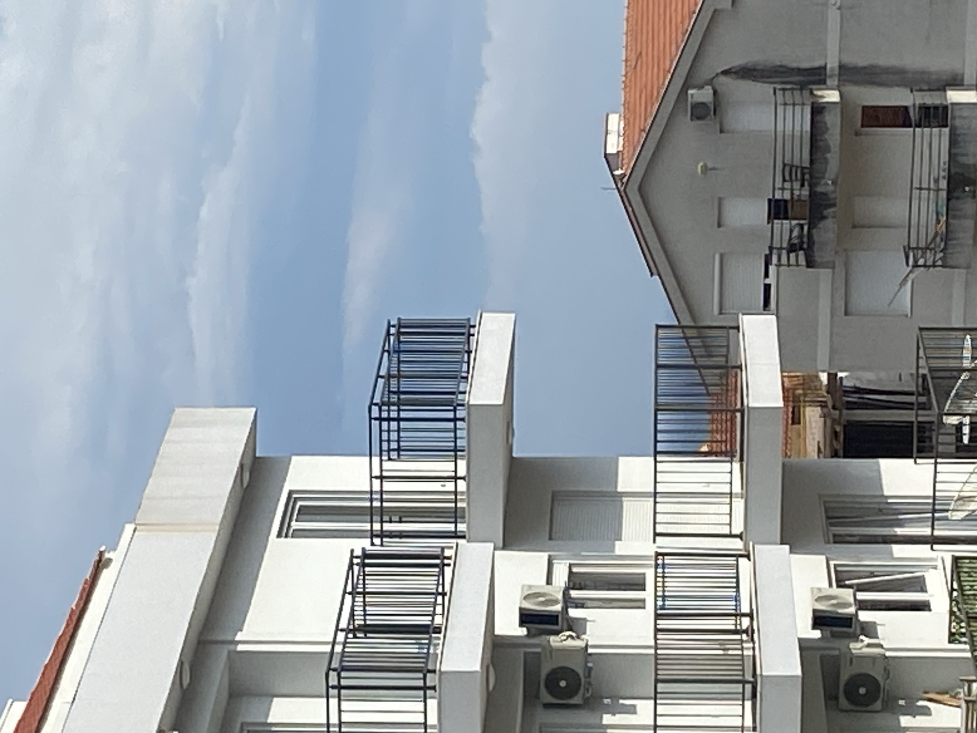 The image displays a vertical shot of two distinct apartment buildings against a clear, bright blue sky with wispy white clouds, typical of an urban setting in Budva, Montenegro. The strong daylight suggests it is midday or early afternoon.
On the right side of the frame, an older building features a reddish-brown tiled roof and light gray walls. Notable dark, vertical stains or water marks extend down from below the roofline. This building includes multiple windows, some with closed white roller shutters, and simple black metal railings, likely for balconies or window guards. An air conditioning unit is visible on its upper facade.
Dominating the left and central portion of the image is a more modern, multi-story white building with a clean, angular design. This structure is adorned with prominent black metal railings, which appear to be part of numerous balconies. Its pristine white walls contrast with the older building. Several windows, some fitted with closed white roller shutters, are visible, along with multiple air conditioning units mounted on its facade, particularly on the lower levels captured in the frame. One window in the mid-section of this building appears to be open.
The scene is devoid of people or discernible activity, focusing solely on the architectural elements. There is no visible text. The overall impression is one of static architecture under clear, sunny weather.
