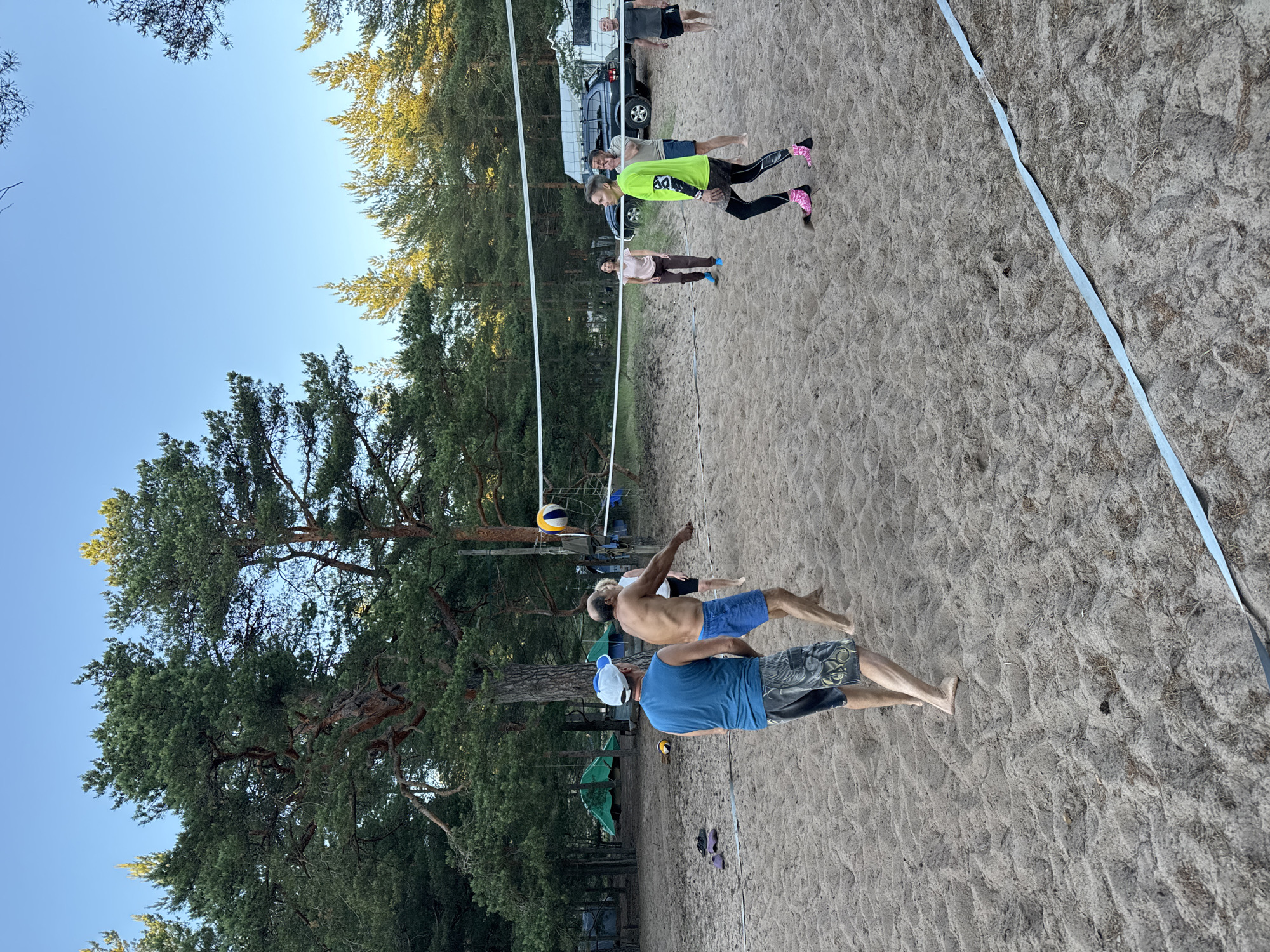 An outdoor daytime scene in Zelenaya Roshcha, Russia, features a volleyball game on a sand court surrounded by pine trees. Six individuals are actively participating in the game. In the foreground, two men are on the near side of the net. The man on the left, wearing a blue sleeveless shirt, a blue baseball cap, and patterned shorts, is facing away from the camera, observing the game. The man on the right, shirtless and wearing blue shorts, is mid-action, with his arm extended as if playing. On the far side of the net, four people are visible. A woman in a light brown t-shirt and dark pants is on the left. To her right, a person in a bright green long-sleeved shirt and black leggings with pink shoes is looking towards the ball. Behind them, two men are standing, one in a grey t-shirt and dark shorts, and the other in a dark t-shirt and dark shorts. A volleyball is in the air above the net. The environment consists of a sandy court with visible sand textures and scattered dried grass. A volleyball net stretches across the court. In the background, dense pine trees form a backdrop, and several green tents are visible among the trees, suggesting a campsite. A dark SUV and a white camper van are parked behind the net, near the trees. The sky is clear and blue.