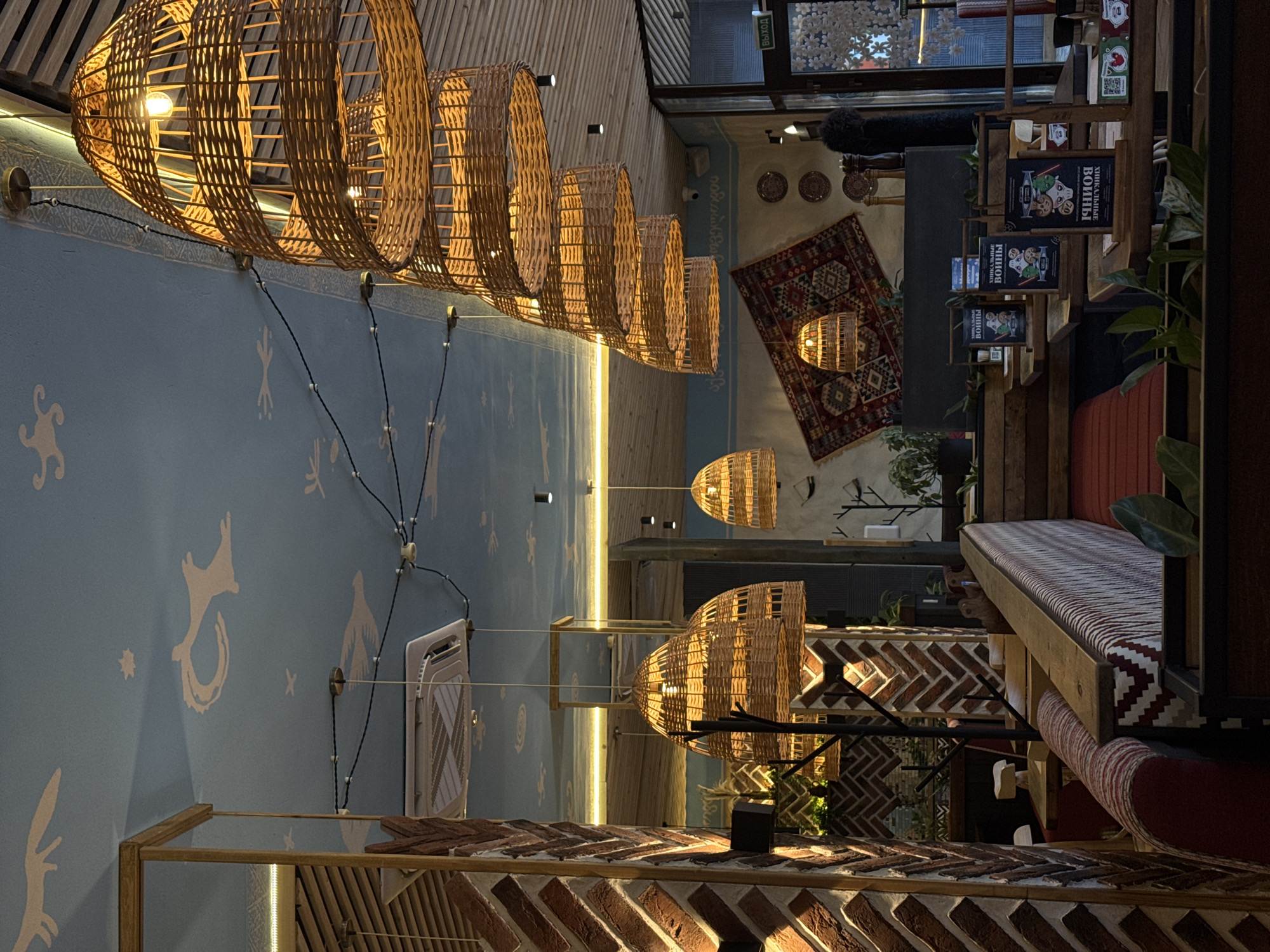 The image depicts the interior of a warmly lit establishment, possibly a cafe or restaurant, with a distinct rustic and cozy ambiance. The scene is dominated by several large, woven lampshades that hang from the ceiling, casting a golden glow. These lamps are intricately made from natural materials, likely rattan or bamboo, and their open weave allows the light to diffuse softly.
The walls are painted a light blue and adorned with whimsical white stencils of animals and abstract shapes, giving the space a playful and artistic character. A patterned rug with geometric designs is laid out on the floor in the background, adding to the eclectic decor. Shelving units displaying books and decorative items are visible, hinting at a place that might also serve as a small bookstore or art space.
Further details include exposed brickwork used in a decorative pattern on some walls and surfaces, contributing to the rustic feel. A cushioned bench with a striped red and white pattern is also present, suggesting seating areas. The lighting suggests it might be evening or an overcast day, as the artificial light is the primary illumination. There's no explicit indication of people engaged in specific activities, but the overall atmosphere suggests a place designed for relaxation and enjoyment. Some visible text on book covers appears to be in Cyrillic script, consistent with a Russian location. The overall impression is one of comfort, warmth, and a carefully curated, bohemian-inspired aesthetic.