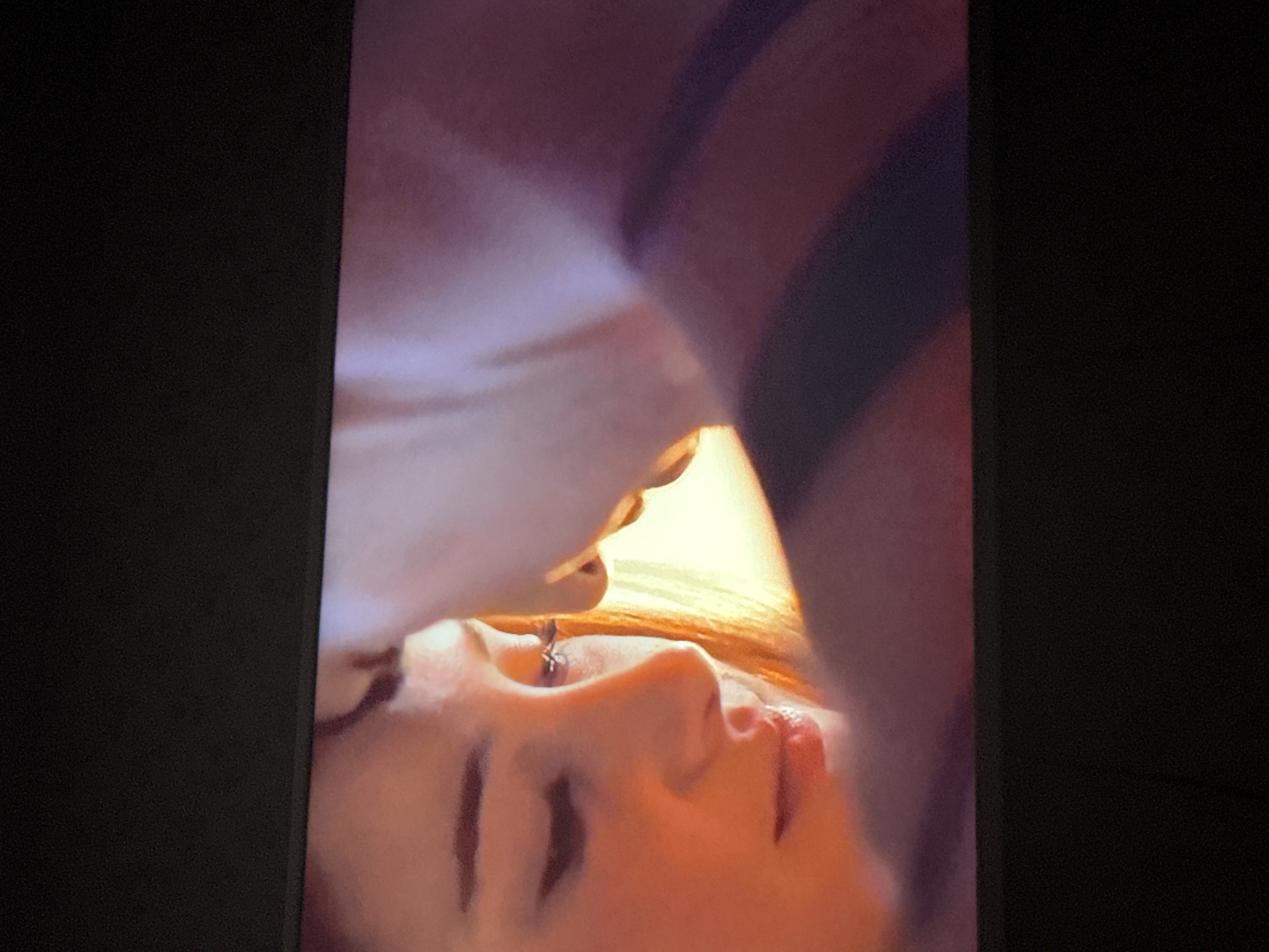The image shows a close-up of a person's face, possibly a woman, in a slightly upward angle. The person is lying down, as suggested by the orientation of the face and hair. The face is illuminated with a warm light, suggesting an indoor environment with artificial lighting. Part of another person's body, perhaps a shoulder or chest, is visible above, indicating a close interaction between the individuals. Details like facial features (nose, lips, eyebrows, and eyelashes) are noticeable. The image appears to be a screen capture or a photo taken of a screen, given the dark borders around the image. There is no information or visual indication to suggest the location is in the city of Mytishchi, Russia.