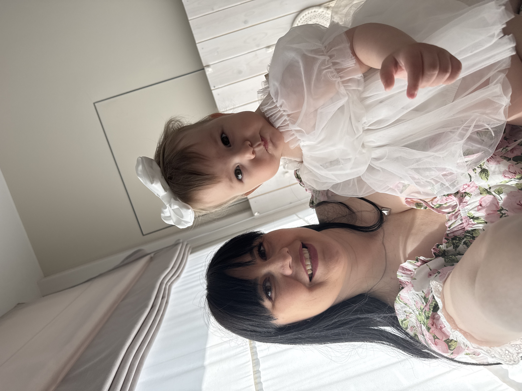 A woman with dark hair and a floral print top is lying down and smiling at the camera, with a baby wearing a white dress and a white bow in her hair positioned above her. They are indoors. The room includes white wooden planks and a white wall. The photo may have been taken in Izhevsk, Russia, but there is no way to know from the image.