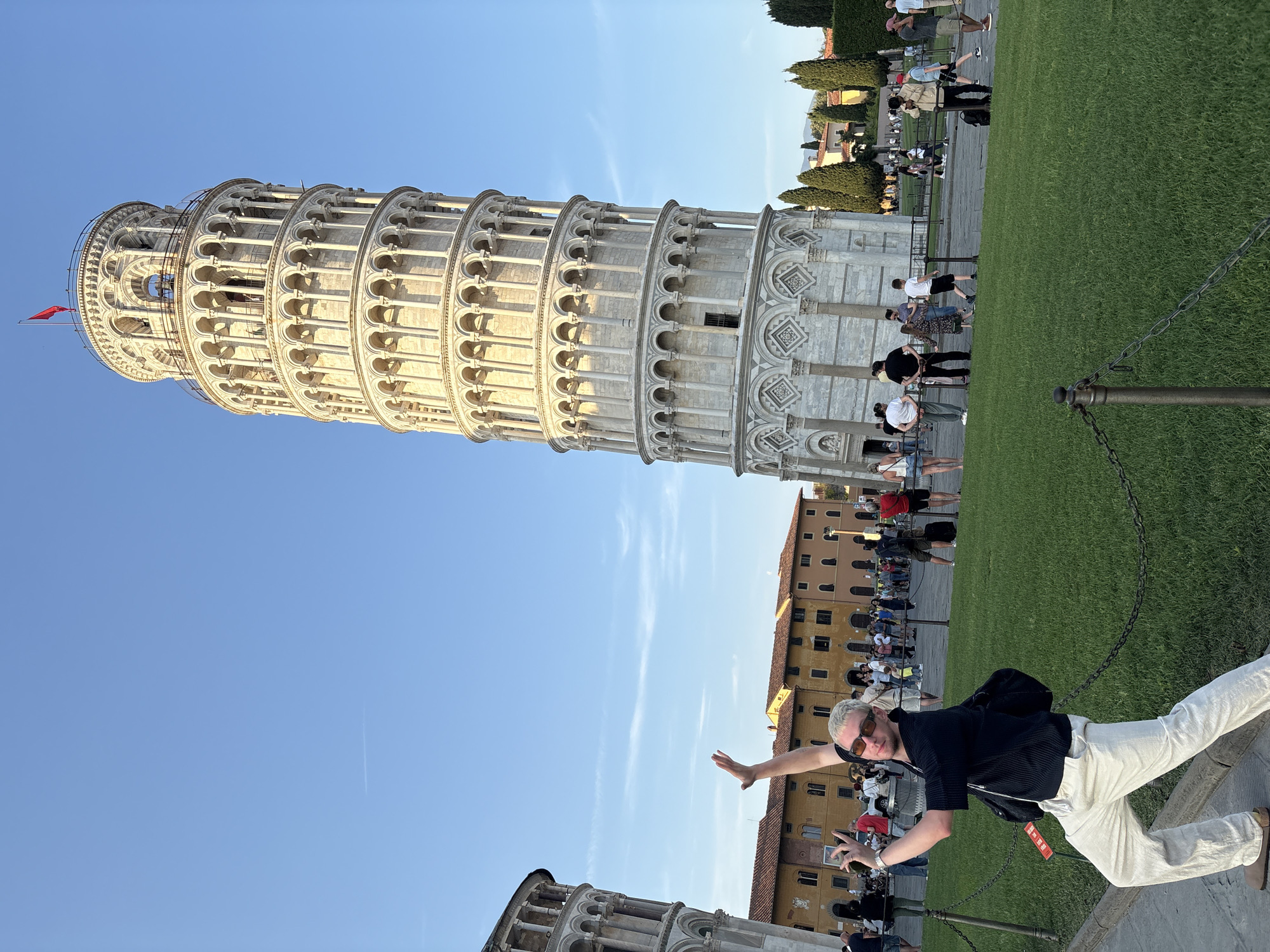 The image captures a vibrant tourist scene at the Leaning Tower of Pisa in Italy, under a clear blue sky. The iconic white marble bell tower, with its distinctive lean and multi-tiered arched structure, dominates the left side of the frame. A small red flag is visible at the very top of the tower.
In the foreground, a man with light-colored hair and sunglasses is actively posing, extending his right arm towards the tower as if to push or hold it upright, a popular tourist photo trope. He is wearing a dark, short-sleeved shirt, light-colored pants, and a black backpack, with one leg extended for balance. He holds what appears to be a phone in his left hand.
The setting includes a neatly maintained green lawn, separated from the viewer by a metal chain barrier. Beyond the lawn, various tourists are scattered, walking, observing, or also posing for pictures. In the background, traditional Italian buildings with terracotta roofs are visible, adding to the historical ambiance. The bright sunlight suggests either morning or late afternoon, casting clear shadows and illuminating the tower. The sky is a cloudless, vibrant blue, with a faint contrail visible in one of the cropped hints. No legible text is visible in the image.