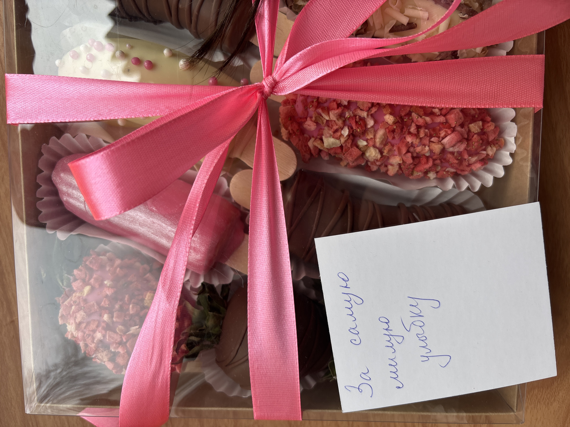 A top-down, medium shot shows a clear plastic box with a pink ribbon tied around it, containing a variety of chocolate-covered strawberries. A card sits next to the box, bearing text in Russian. The background appears to be a wooden surface. It is likely located in Perm, Russia.