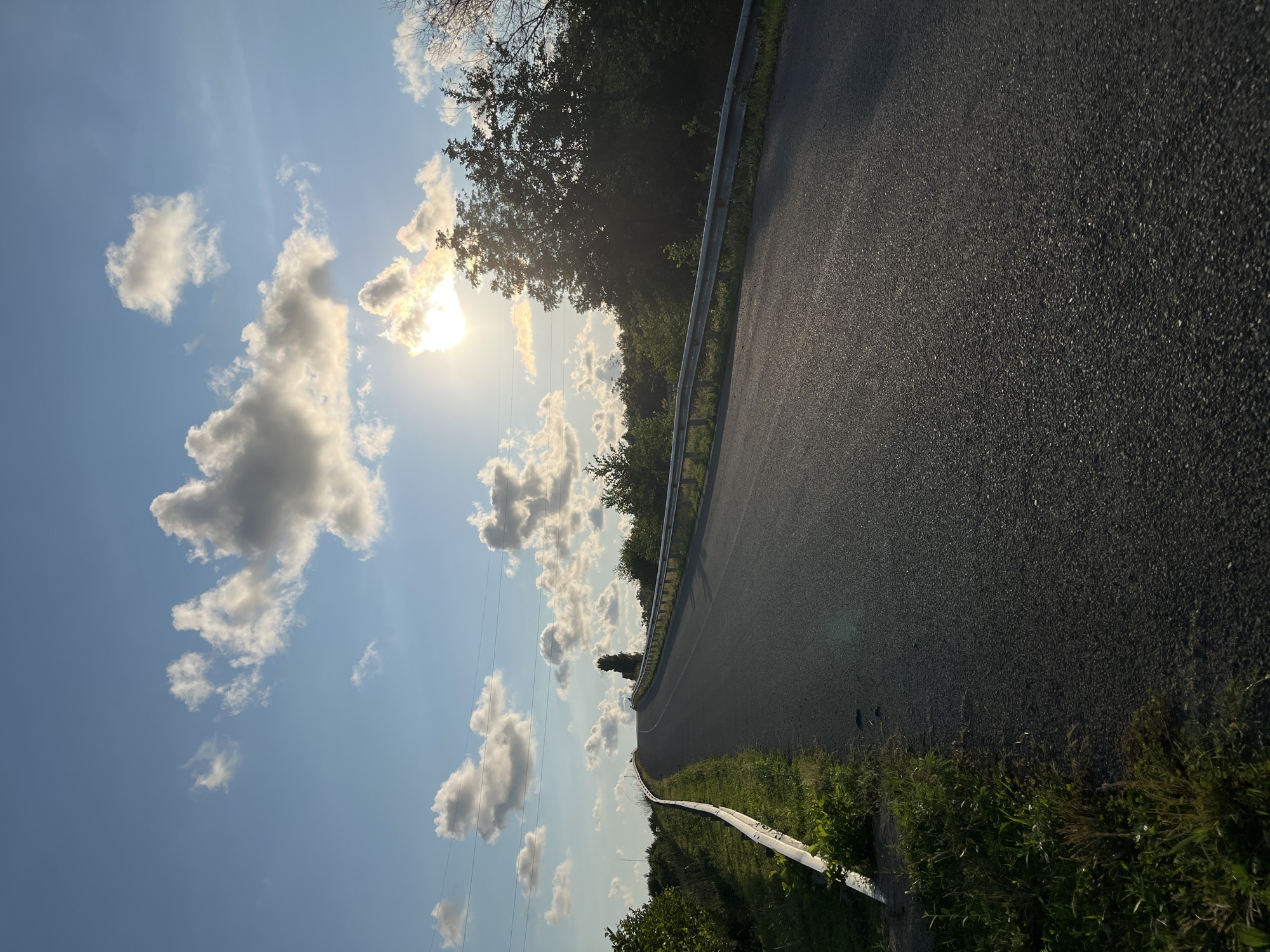 The image captures an upward-sloping asphalt road winding through a rural landscape under a bright, partly cloudy sky. The sun is visible, partially obscured by clouds, casting a warm glow. On the left side of the road, a white guardrail runs along a grassy verge. On the right, a dense line of trees borders the road, with some lighter foliage visible in the background. Utility poles and wires are present in the sky. The scene appears to be a serene, open area, likely outside of a major urban center. Based on the context provided, the location is identified as the city of Ureki, Georgia, suggesting this is a road leading into or out of the town, possibly a scenic route. No people are visible in the image, and no specific actions or interactions are depicted. The environment is characterized by the road, vegetation, sky, and sun.