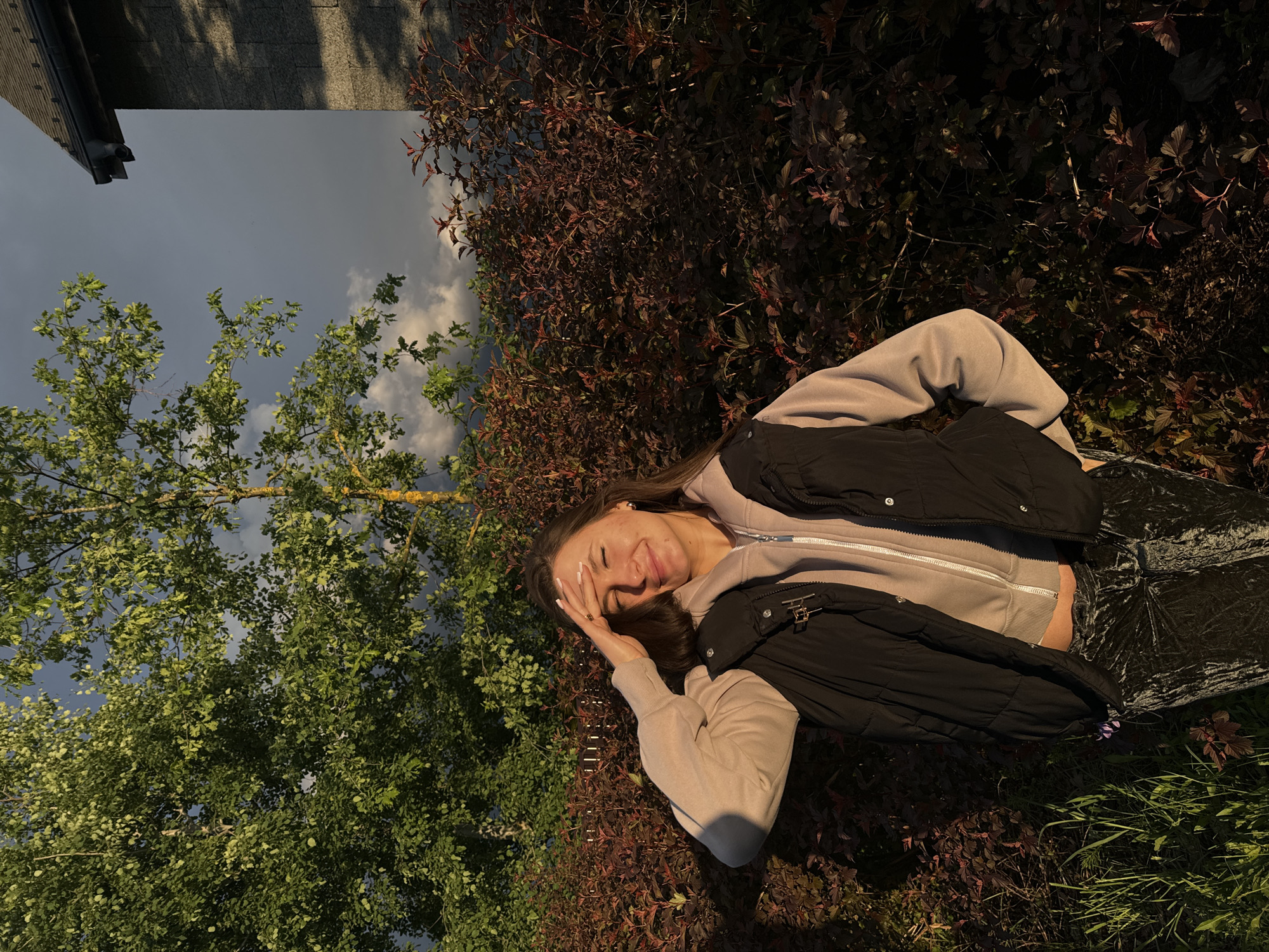 A young woman lies in a grassy area bordered by shrubs and a tree, in what appears to be Vysokinichi, Russia. She is wearing a light-colored hoodie with a black vest and dark pants. Her face is visible, and she has one hand raised near her forehead. Overcast sky visible above the trees. A portion of a building's roof and side are visible in the background. The scene is lit by soft, diffuse sunlight.