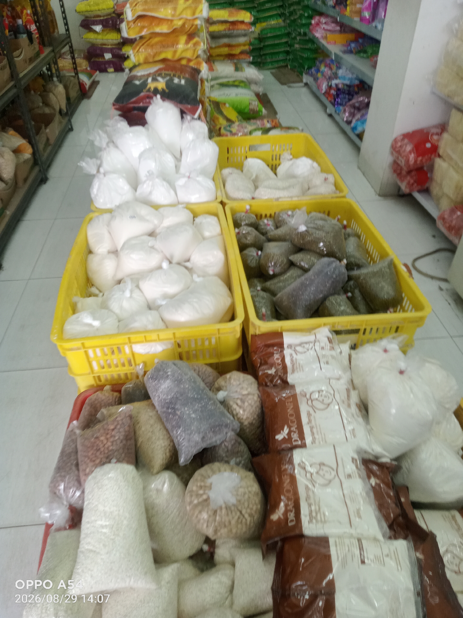 The image shows a store interior with shelves stocked with various food items, primarily packaged grains and powders. In the foreground, several large, clear plastic bags are filled with white granular material, likely rice or flour. To the right, there are also several brown and white bags with text that appears to be in an Asian language, possibly featuring a logo with a dragon. Yellow plastic crates are filled with more bagged food items. In the background, shelves are stacked with more large bags of food products, some appearing to be rice sacks with colorful designs. The lighting suggests an indoor environment, typical of a retail space. The scene is devoid of people. The timestamp in the lower-left corner, "2026/08/29 14:07," and the "OPPO A54" text indicate the photo was taken with an OPPO A54 phone on August 29, 2026, at 2:07 PM. While the image itself does not contain explicit geographical markers, the presence of a timestamp from a specific date and time suggests this is a photograph taken at a particular moment.