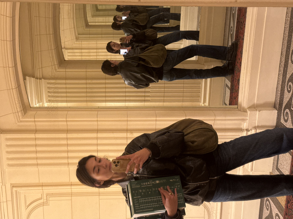 A female individual, appearing East Asian with short dark hair, is depicted taking a selfie in a grand, ornate interior hallway. She stands facing the camera, holding a light-colored smartphone in her right hand, angled for the self-portrait. In her left arm, she carries a stack of two large, green bound books or binders. She wears a dark, possibly leather, bomber jacket, dark blue wide-leg jeans, and dark shoes. A large, olive-green, soft fabric shoulder bag is draped over her left shoulder.
The environment is characterized by classical architecture, strongly suggesting the **Palais Garnier (Opéra National de Paris)** in Paris, France. The hallway features cream-colored stone walls adorned with fluted pilasters, decorative moldings, and cornices. On the right, a sequence of large, rectangular mirrors with arched tops reflects the individual and the hallway, creating an infinite recession of identical images. The floor in the foreground shows a grey and white patterned mosaic, while the reflected sections reveal a dark red and patterned oriental-style rug covering the floor. The scene is evenly lit by warm interior lighting.