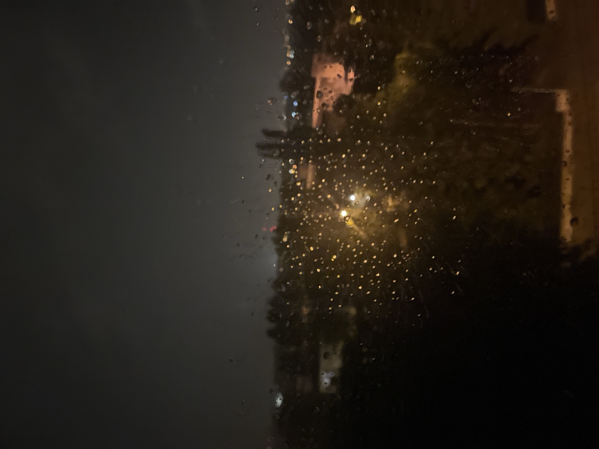 The image is a nighttime view through a rain-streaked window, looking out at a dimly lit cityscape. The foreground is dominated by dark, blurry shapes, likely trees or buildings, with numerous water droplets scattered across the glass, creating bokeh effects with the distant lights. In the mid-ground, faint outlines of buildings are visible, illuminated by scattered streetlights and internal lights. The background shows a dark, cloudy sky with a subtle glow from the city. The overall impression is one of a quiet, possibly rainy night in an urban or suburban environment. No people or specific actions are discernible.