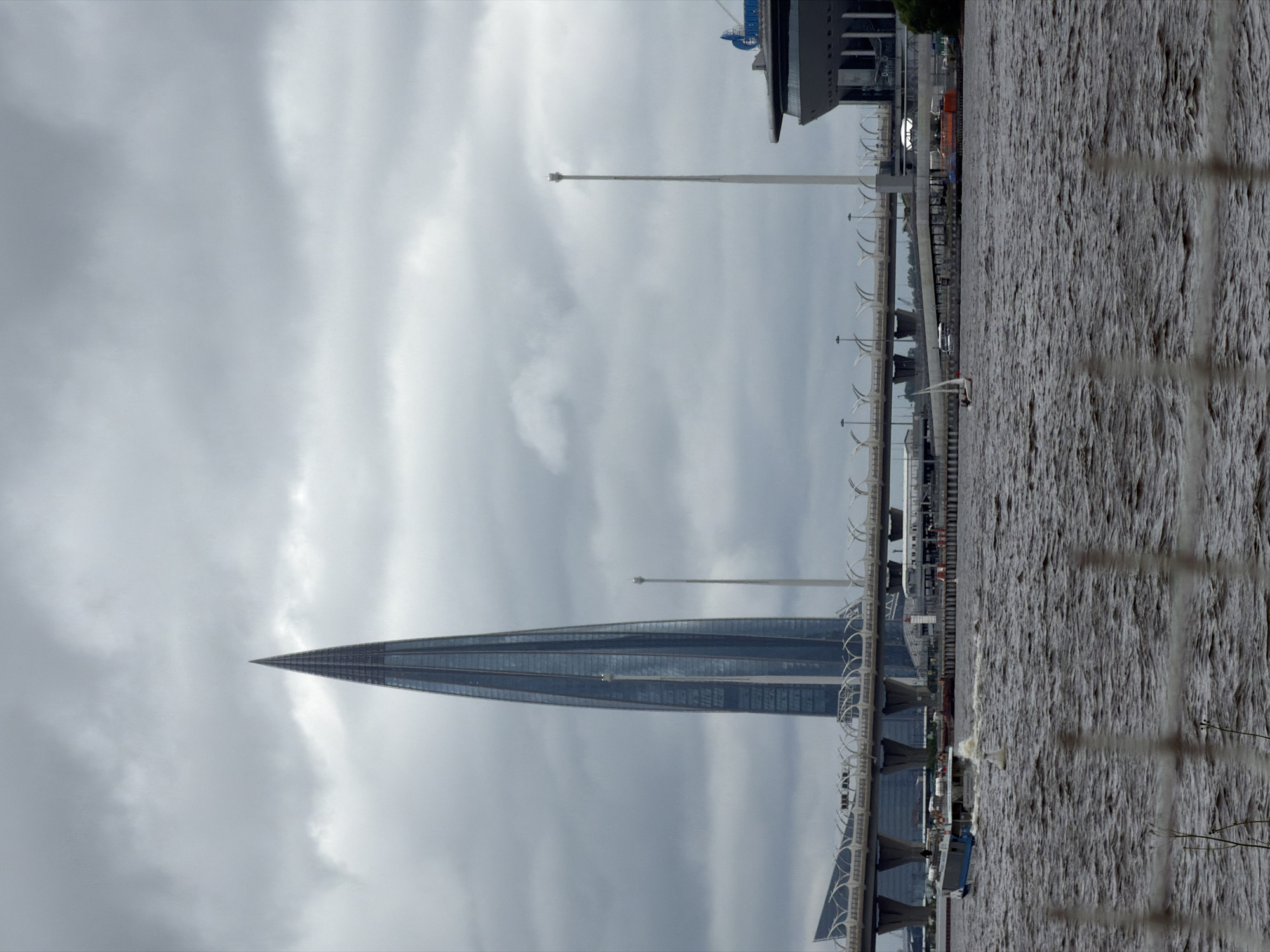 The image depicts a cloudy, overcast day in Saint Petersburg, Russia, with a prominent view of the Lakhta Center skyscraper and the Western High-Speed Diameter bridge. In the foreground, a body of water, likely the Gulf of Finland, is choppy with visible waves. A white fence or railing is out of focus in the very front of the image.
Dominating the left side of the frame is the Lakhta Center, a modern skyscraper with a distinctive spiraling, pointed design, clad in glass. A bridge, part of the Western High-Speed Diameter, spans across the water in front of the skyscraper, supported by large concrete pillars. The bridge has an intricate, arched railing system.
To the right of the Lakhta Center, two tall, slender white poles stand vertically, one appearing taller than the other, possibly lighting fixtures or communication towers. Further to the right, a portion of a large, modern stadium or arena building is visible, with a dark, curved facade and blue architectural elements.
On the water, a ferry or passenger boat is moving from left to right, creating a wake. Further out, a small white sailboat is visible with its sail up. Several other boats and some industrial structures are present along the distant shoreline. The overall impression is of a large urban and infrastructural landscape under a moody sky.