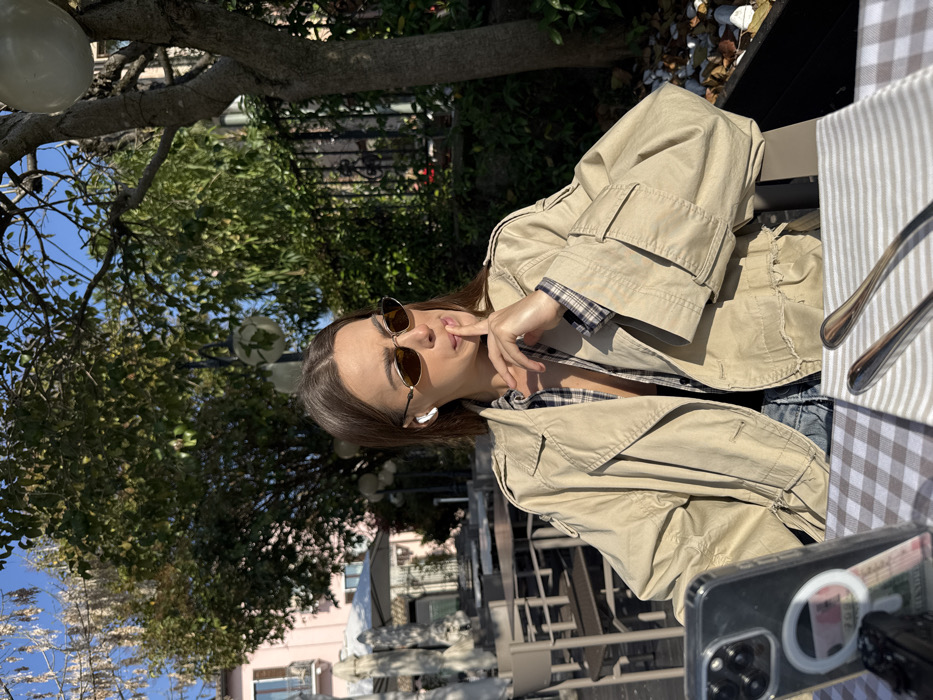 The image depicts an outdoor dining scene at a café or restaurant patio in Sirmione, Italy, under clear blue skies and bright sunlight. An adult female, with long dark hair, is seated at a table in the foreground. She wears oval-framed sunglasses, a white wireless earbud in her left ear, a beige trench coat, and a plaid shirt. Her right index finger is touching her lips, and she looks to her right. The table is covered with a white and brown checkered tablecloth, featuring a striped white napkin and two pieces of silverware. A dark-colored smartphone in a clear case, positioned upright on the table's left, displays a MagSafe ring and currency. The background includes green foliage from trees, a dark metal fence, several empty outdoor dining tables with chairs, and a light pink building. White spherical decorative lights are visible among the tree branches.