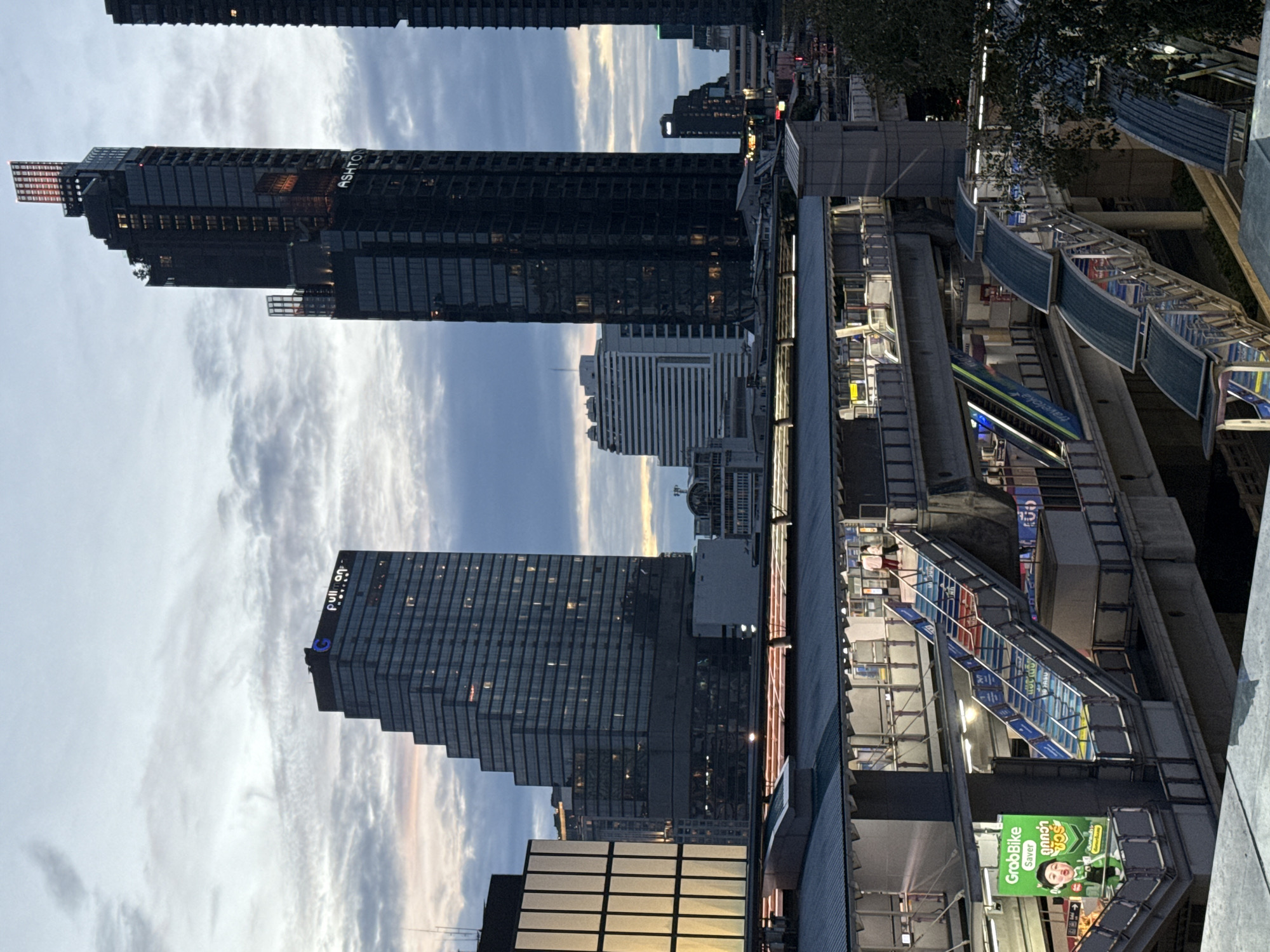 This image depicts a twilight view of a modern urban scene in Bangkok, Thailand, focusing on a multi-level transportation hub and surrounding skyscrapers. The foreground features a skywalk and elevated walkways with a prominent green advertisement for "GrabBike Saver" on the left. Staircases and escalators are visible, some illuminated with colorful lights. People are seen walking on the walkways, suggesting active use of the transit system.
In the midground and background, several tall buildings dominate the skyline. To the left, a stepped, glass-faced skyscraper displays the "Pullman Hotels" logo. Adjacent to it is a darker, slender skyscraper with illuminated windows and the "ASHTON" branding near its top. Further into the background, other buildings of varying architectural styles, including a horizontally paneled white building, contribute to the dense cityscape. The sky is filled with dramatic clouds, indicative of the transitional light of dusk or dawn. The overall impression is one of a bustling, contemporary metropolis.