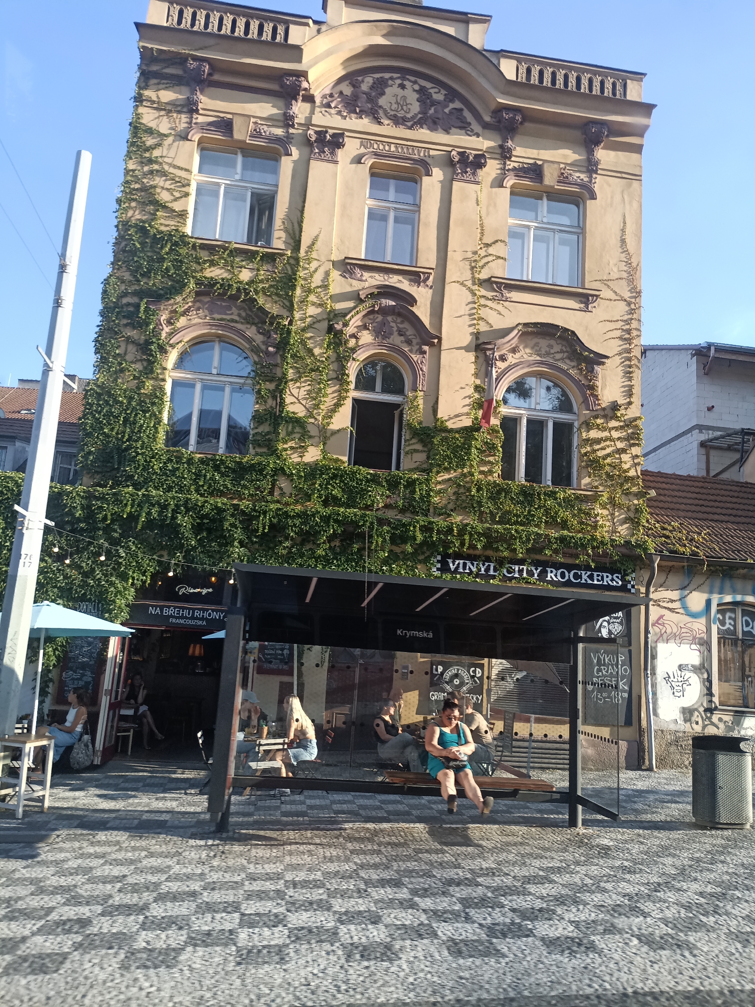The image shows a building with ivy growing on its facade, in Prague, Czechia. In the foreground, there is a bus stop with a shelter. Several people are sitting on the benches at the bus stop or at tables outside a cafe. The building has architectural details and several windows. The street is paved with cobblestones. There are signs on the building and at the bus stop, one of which reads "VINYL CITY ROCKERS."