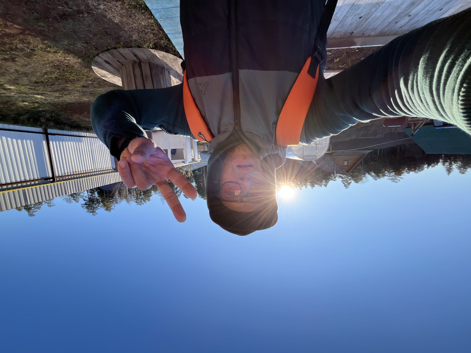 An outdoor selfie captures a person making a peace sign under a clear, bright blue sky in Chaikovsky, Russia. The image is taken from a slightly low angle, showing the person from the chest up, with their head tilted slightly. They are wearing glasses, a dark hooded top, and a two-toned jacket (gray and dark blue/black) featuring bright orange straps or accents over the shoulders. A small, stylized logo, resembling a "W" or an animal head, is visible on the gray section of the jacket, and partial text, possibly "COLO," is on the left sleeve. The individual's left hand is raised, forming a peace sign.
The sun is bright and low in the sky, creating a prominent lens flare around the person's head, partially obscuring their face and casting strong highlights on their right side. In the background, a rural or semi-rural setting unfolds. Distant evergreen trees line the horizon beneath the expansive blue sky. Several structures are visible, including a small wooden cabin or shed with a dark roof, a white corrugated metal fence, and a greenhouse-like tunnel structure. A large wooden cable spool, repurposed as outdoor furniture, is in the mid-ground. The ground visible near the wooden spool shows sparse grass and dirt. The scene depicts a sunny day, likely morning or late afternoon, given the sun's angle and intensity.