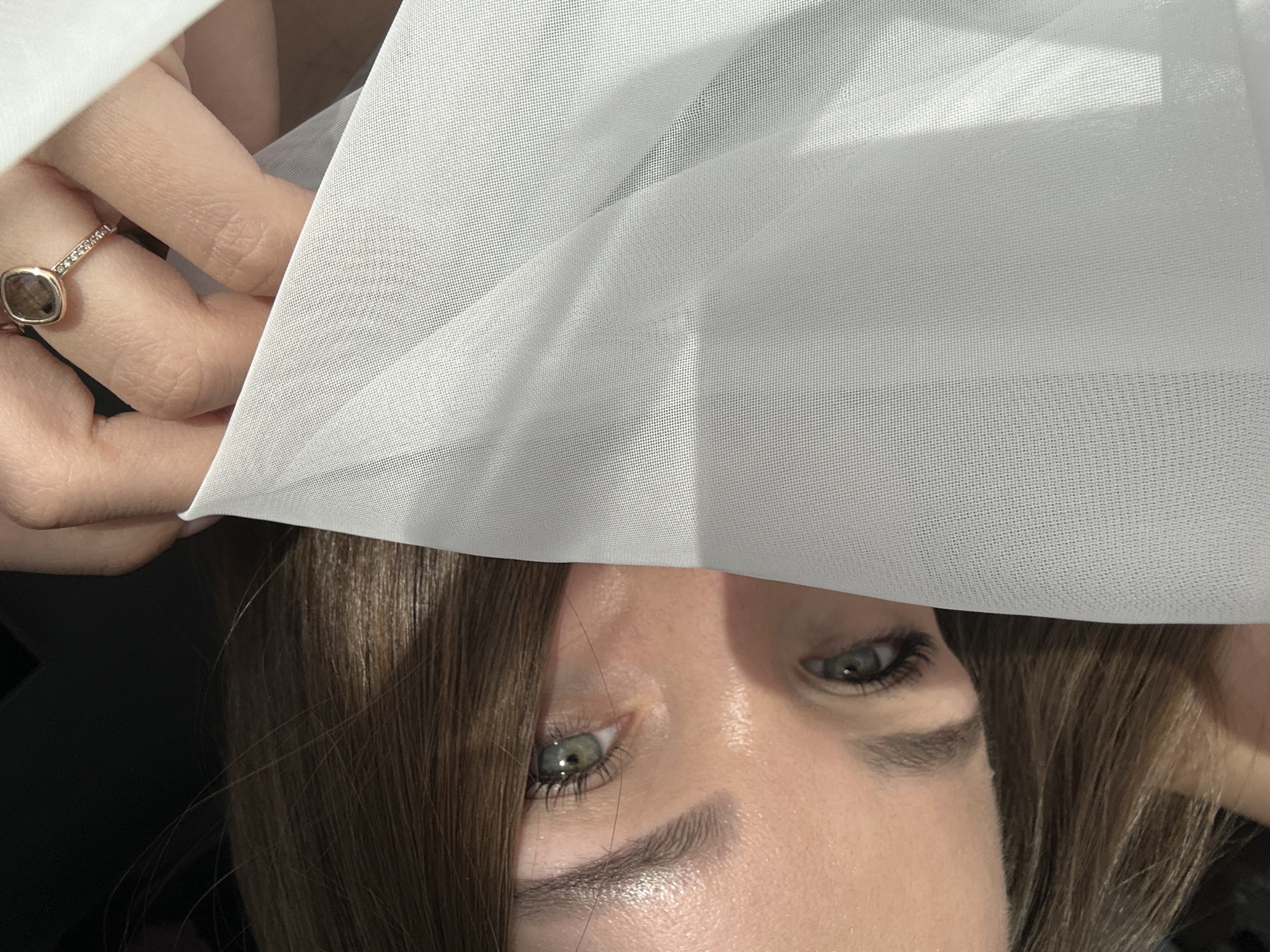 An image, captured in Chaikovsky, Russia, presents a close-up, oblique view of a female subject partially obscured by sheer white fabric. The subject's visible features include her left eye and the upper portion of her right eye, both with green/hazel irises and dark eyelashes. Defined dark brown eyebrows are present. Brown hair is visible on the left and lower sections of the frame. A right hand is positioned in the upper right quadrant, holding the finely-meshed white fabric. A gold-toned ring, featuring a cushion-cut dark stone and small clear stones on the band, is worn on the ring finger of the visible hand. The fabric covers the lower right area of the subject's face and extends across the right side of the image. Strong direct light illuminates the subject's skin and the fabric, while the background behind the subject's hair in the upper left is dark and undifferentiated.